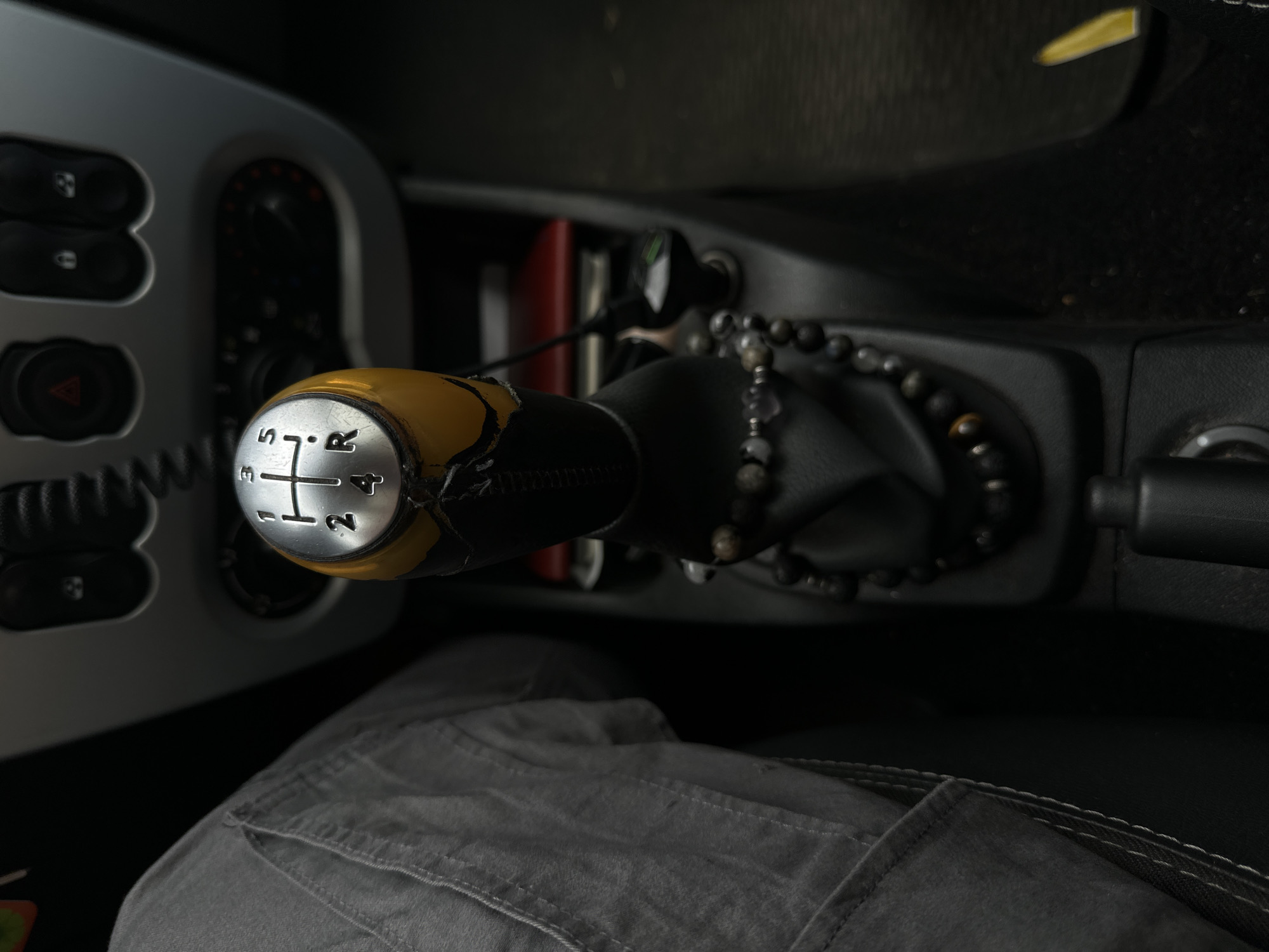 A close-up, eye-level shot focuses on a yellow and silver gear shift knob in a vehicle. The gear pattern is visible, showing positions 1, 3, 5, 2, 4, and R. The knob has some wear and tear, with exposed yellow material beneath the silver top. A string of dark beads is wrapped around the base of the gear shift. To the left, a portion of gray pants is visible, suggesting someone is seated in the driver's seat. The background shows a section of the car's dashboard with various buttons and controls. The lighting is dim, creating a moody atmosphere. The scene appears to be located within a vehicle, potentially parked, in the Бараўлянскі сельскі Савет region of Belarus.