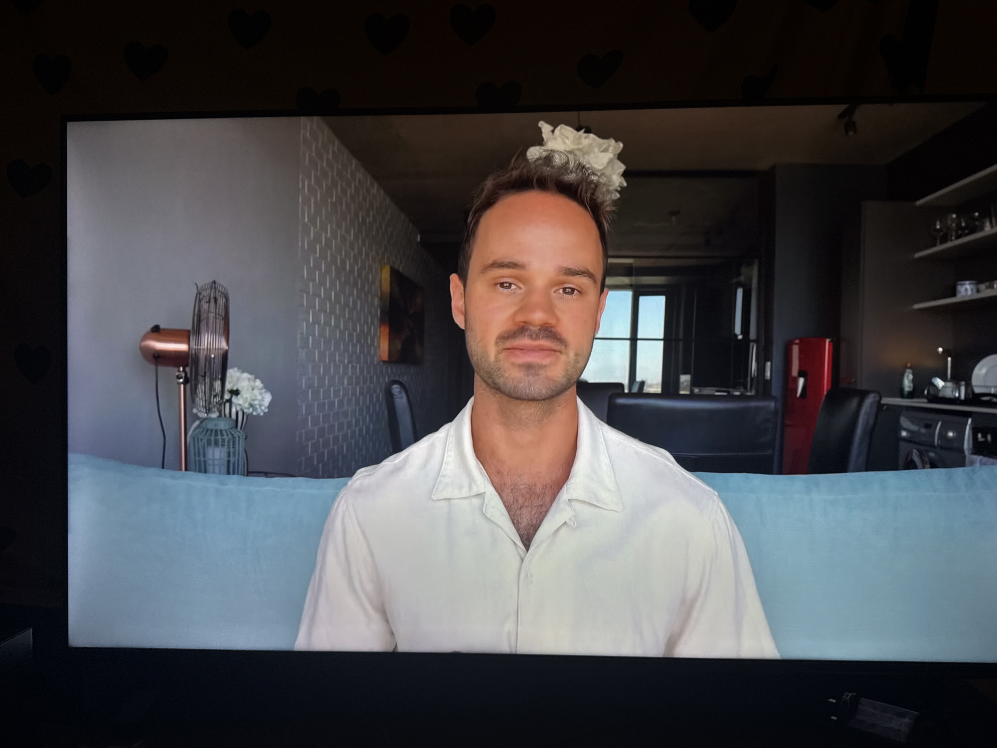 A man with short brown hair and a light stubble is seated on a light blue couch, facing the camera. He is wearing an unbuttoned, short-sleeved white shirt. A white, fluffy object is perched on top of his head. Behind him, a room is visible, featuring a patterned wall on the left, a vintage fan with a copper accent, and a vase with white flowers. The background also includes a kitchen area with dark cabinets, white shelves, a red refrigerator, and windows. The scene appears to be indoors, likely a residential apartment. There is no indication of the location being in Cheltenham, United Kingdom.