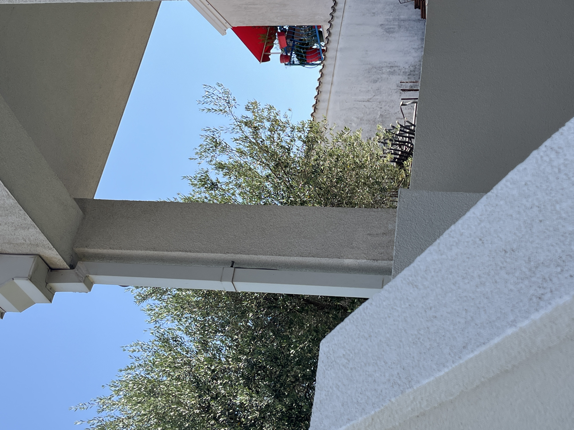 The image depicts an outdoor scene with a bright blue sky. In the foreground, a textured white structure, possibly a balcony railing or stair banister, occupies the lower left portion of the frame. Behind this, a light gray column supports an overhang. To the left of the column, a large, leafy green tree, likely an olive tree, fills the mid-ground. Further back, a white building with a red awning is visible. The awning extends over a small balcony area where red planters and other objects can be seen. A portion of a textured white wall with a terracotta tile roofline separates the mid-ground from the background building. The overall impression is one of a sunny day in a residential area, potentially in Budva, Montenegro, given the architecture and vegetation.