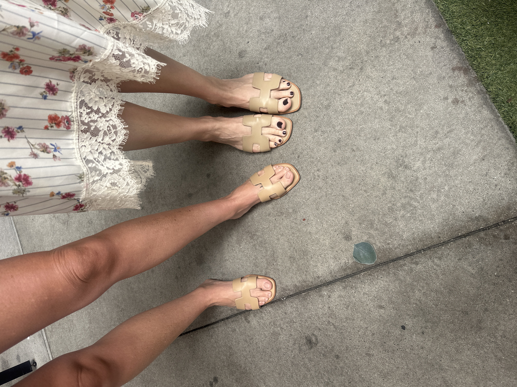 The image shows the lower portions of two people standing on a concrete sidewalk. The person on the left wears a floral dress with a white lace hem and beige slide sandals. The person on the right also wears beige slide sandals. A fallen eucalyptus leaf lies on the sidewalk near the feet of the person on the right. The setting appears to be an urban area, potentially near a commercial building, as indicated by the small patch of artificial grass in the background.