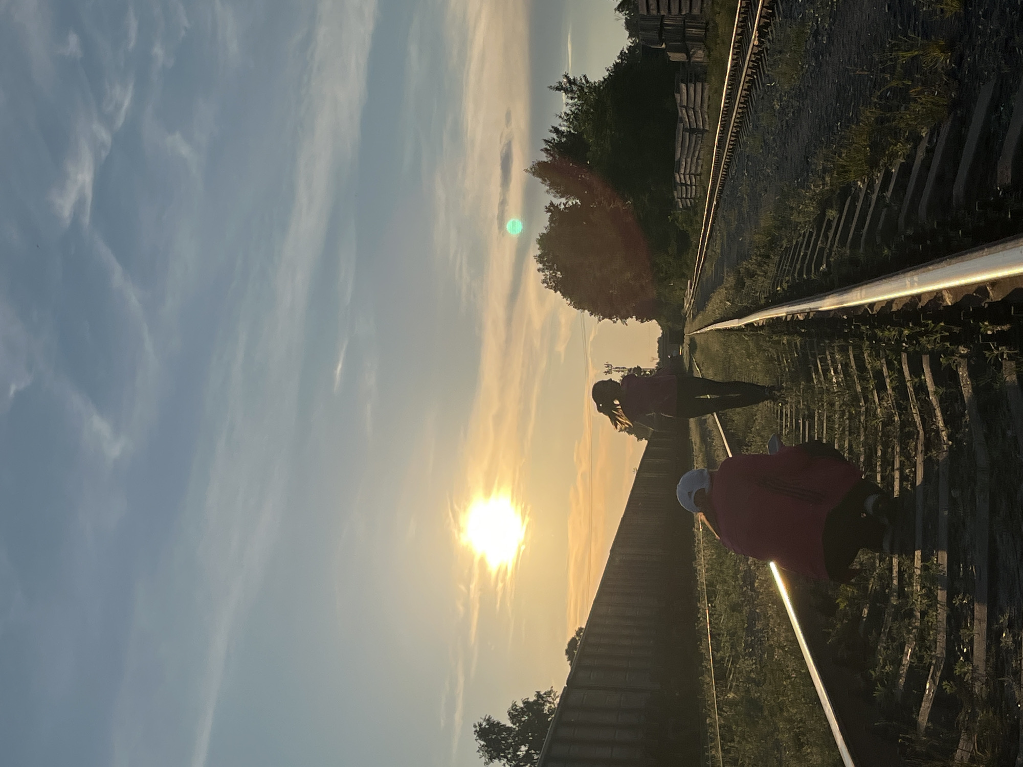 The image depicts two young females standing on a railway track during what appears to be sunset. The sun is low in the sky, casting a warm glow and creating lens flare effects. The girl in the foreground is squatting, wearing a red jacket and a white cap, and facing away from the camera. The second girl is walking away from the camera further down the track, wearing a pink top and dark pants. The railway line stretches into the distance, flanked by overgrown grass and ballast. On the left, a tall, dark fence runs parallel to the tracks, possibly a boundary wall. On the right, a dense line of trees and some stacked materials, possibly construction supplies, are visible. The sky is a gradient of blue and orange, with scattered clouds. The scene is set in a semi-industrial or rural area, consistent with the outskirts of a city like Chaikovsky, Russia, where railway infrastructure is common. The lighting suggests it is late afternoon or early evening.