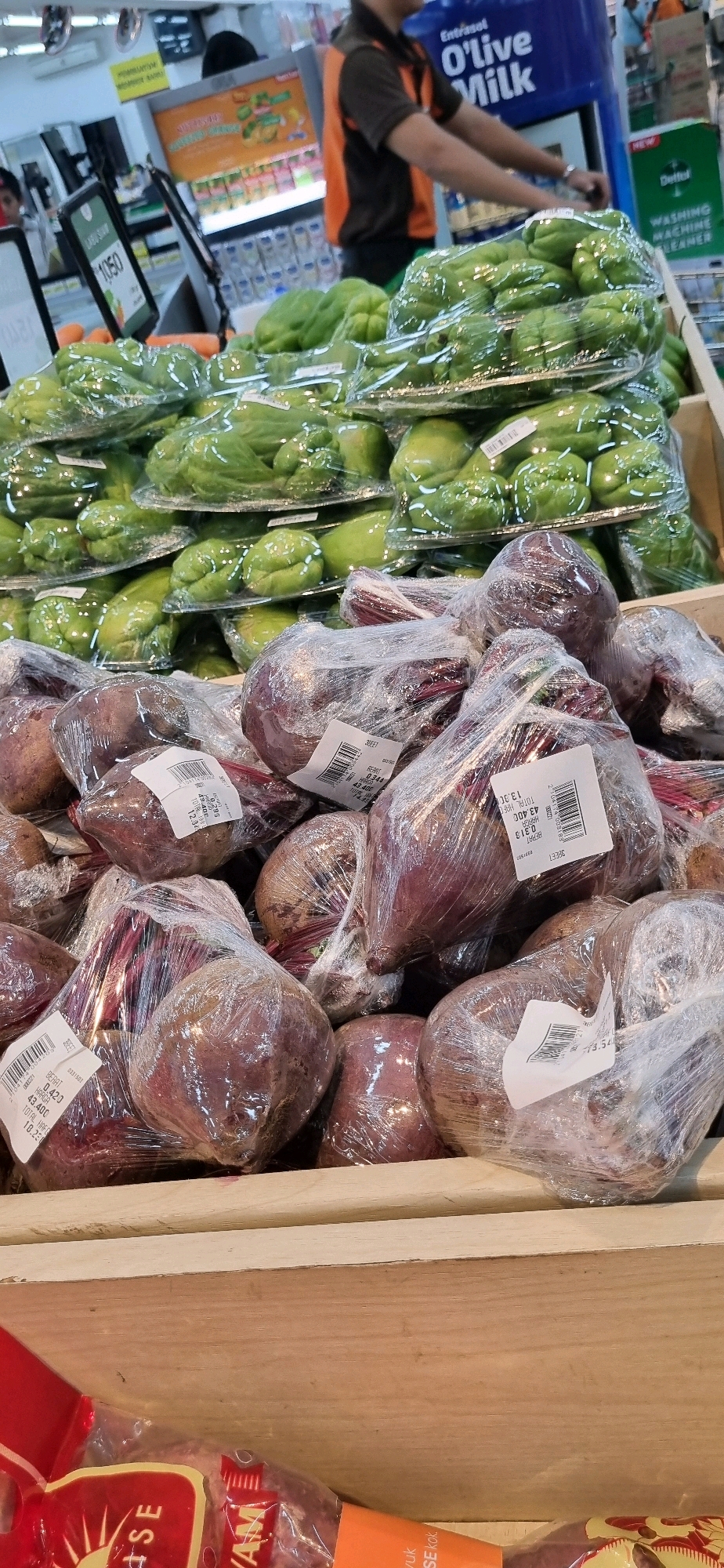 This is a photograph of produce on display in a supermarket in Denpasar, Indonesia. In the foreground, several beetroots are individually wrapped in clear plastic. Behind them, piles of chayote squash, also wrapped in clear plastic, are stacked on shelves. A person wearing an orange and black uniform is partially visible in the background, pushing a shopping cart. The supermarket environment is indicated by shelving units and signage, some of which appear to be in Indonesian. The overall scene depicts a commercial setting for the sale of fresh produce.