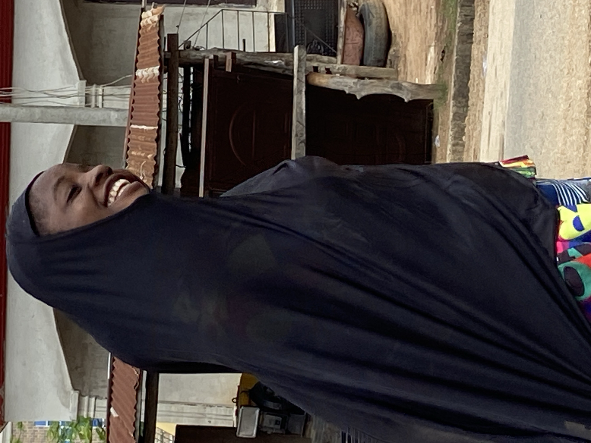 This image captures a joyful moment featuring a person, likely a young female, with dark skin, smiling broadly and revealing a full set of white teeth. Her head is tilted slightly, and her face expresses clear amusement or happiness. She is wearing a dark, possibly black, head covering or veil that drapes over her body, covering most of her clothing except for a small section of a vibrant, colorful, patterned garment visible on her lower right side.
The setting appears to be an outdoor, urban or semi-urban environment in Mararaba, Nigeria, illuminated by daylight. The lighting is soft and diffused, suggesting an overcast day or indirect sunlight, as there are no harsh shadows.
In the background, various weathered structures are visible. To the viewer's left, there are reddish-brown corrugated metal sheets, possibly part of a roof or wall, alongside a light-colored concrete wall and some visible wires. To the viewer's right and behind the person, a dark door or gate with a circular embossed design is partially seen, flanked by wooden beams. Further into the background, a dark vehicle tire leans against a structure. The combination of materials like corrugated metal, concrete, and exposed wires indicates a modest, everyday street scene. There is no visible text in the image.