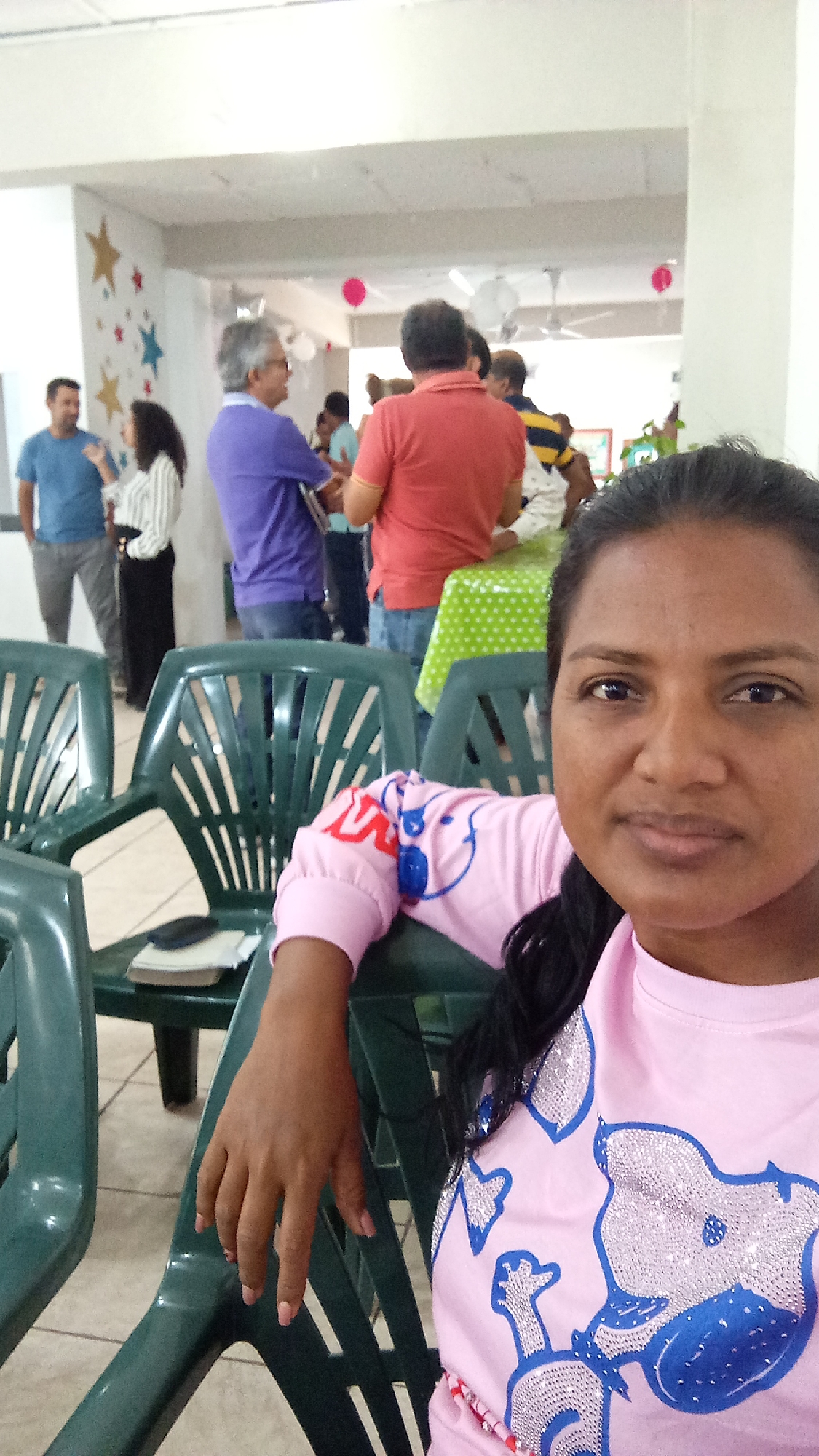 The image shows a woman in a pink long-sleeved shirt taking a selfie. She is sitting in a green plastic chair. In the background, there is a group of people standing and talking. There are stars on the wall and balloons hanging from the ceiling, suggesting a party or gathering. A table covered with a green and white polka-dot tablecloth is visible in the background. The location is the city of Unknown, Unknown.