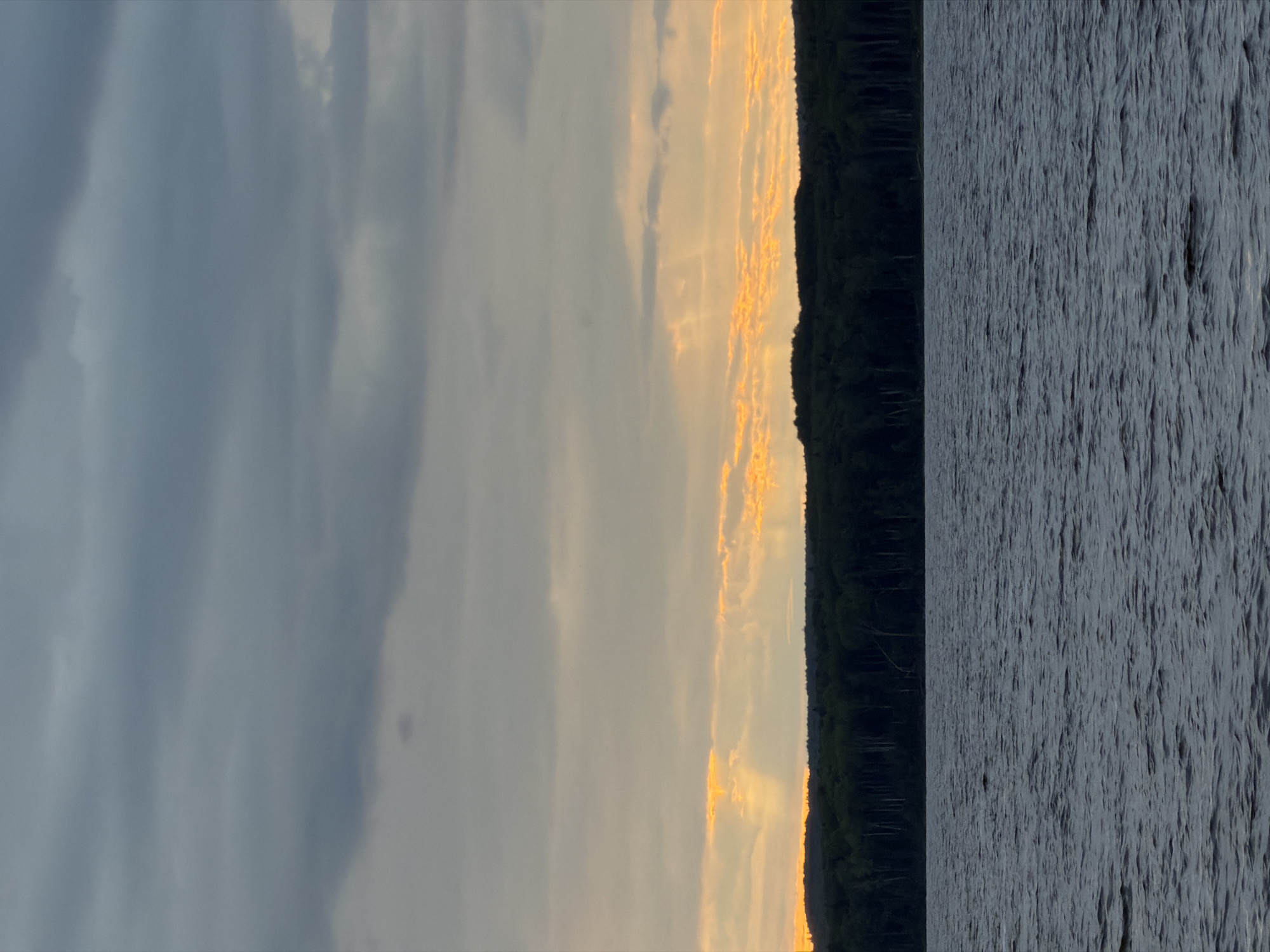 This vertical image depicts a natural landscape scene at what appears to be either sunset or sunrise, located in Chaikovsky, Russia. The sky occupies the upper two-thirds of the frame, characterized by layers of clouds. The uppermost clouds are dark grey and dense, transitioning to lighter grey clouds below. A prominent band of intense orange and yellow light, indicative of crepuscular rays, pierces through the lower cloud layers just above the horizon, casting distinct shafts of light downwards. Directly beneath the illuminated sky, a dense, dark green forest forms a silhouetted horizon line, its undulating top edge suggesting varied tree heights. The lower third of the image is dominated by a dark blue-grey body of water, its surface exhibiting numerous ripples and small waves, suggesting movement from wind or current. There are no people, identifiable structures, or objects of human origin visible within the scene. The overall impression is one of a natural, serene environment under dramatic sky conditions.