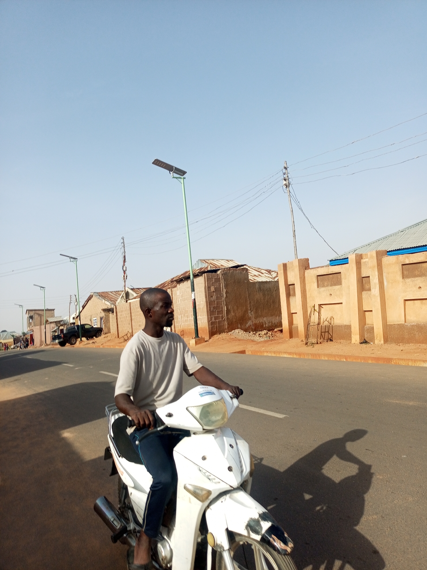 A man is riding a white motorcycle on a paved road in Tumfure, Nigeria. He is wearing a gray shirt and dark pants. The road is lined with buildings and streetlights. Power lines run overhead. A black truck is parked in the distance. The sky is clear. The lighting suggests daytime.