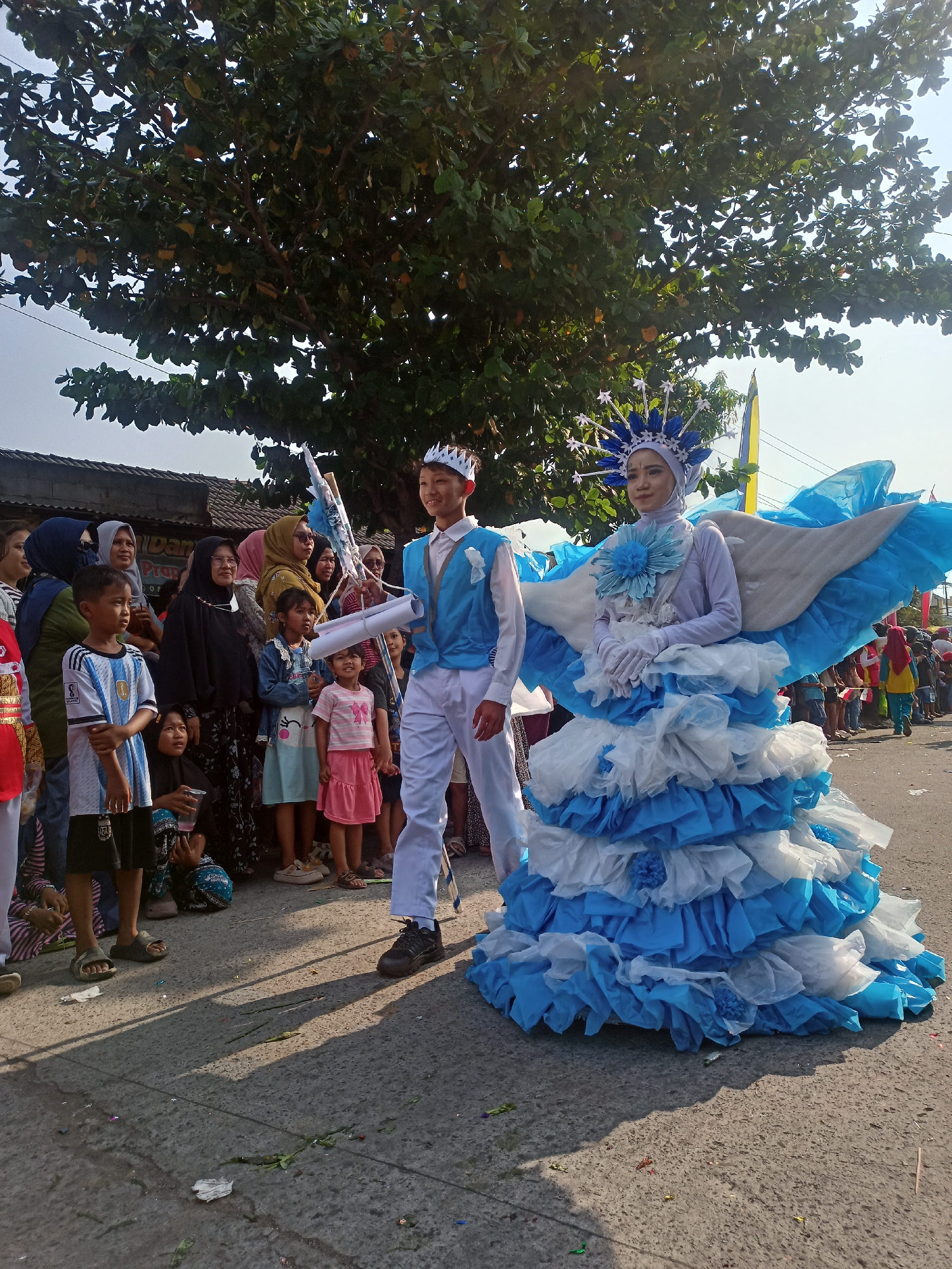 A street parade is underway in Saren, Indonesia, with a large crowd gathered to watch. In the foreground, a young man and woman are participating in the parade, dressed in elaborate costumes. The man wears a light blue suit and a white crown. The woman is adorned in a striking blue and white costume that resembles wings and a layered skirt, also wearing a detailed headpiece. Behind them, a diverse group of spectators, including women in hijabs and children, are observing the procession. The background shows a street lined with buildings and large trees, suggesting a residential or commercial area within the city. The sky is clear and bright, indicating a sunny day.