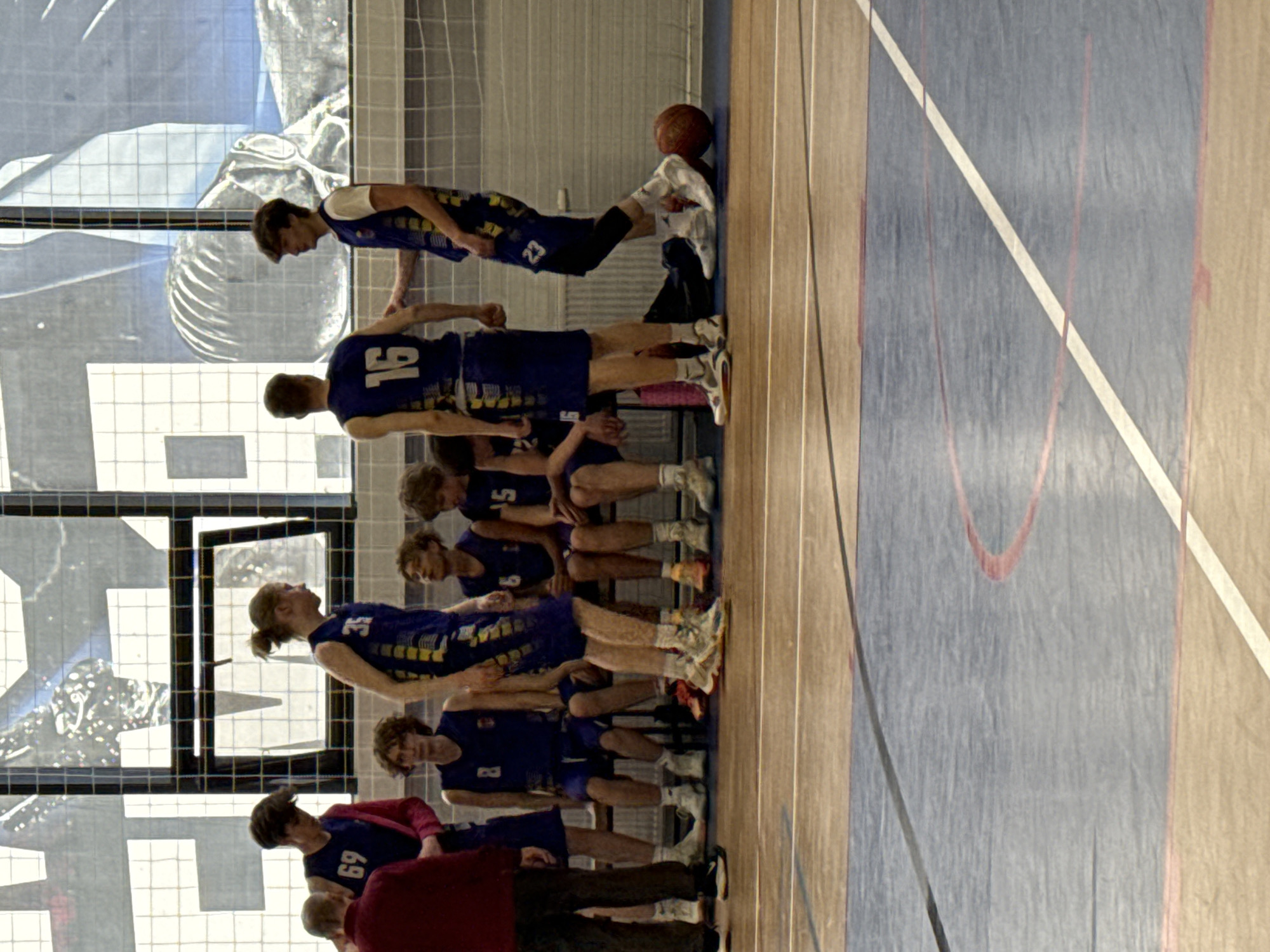 A youth basketball team poses along the edge of a basketball court in what appears to be a gymnasium. The team, dressed in blue and yellow basketball uniforms, consists of about 10-12 members, lined up in a slightly staggered row. The player at the end of the line near the wall is holding a basketball on top of a towel, seemingly using his leg to prop it up. Some adults, presumably coaches or guardians, are standing near the back of the group. The basketball court has a traditional design with wooden floor panels and painted lines. In the background, a large window allows natural light into the gym. The location is consistent with an indoor sporting facility within Votkinsk, Russia.