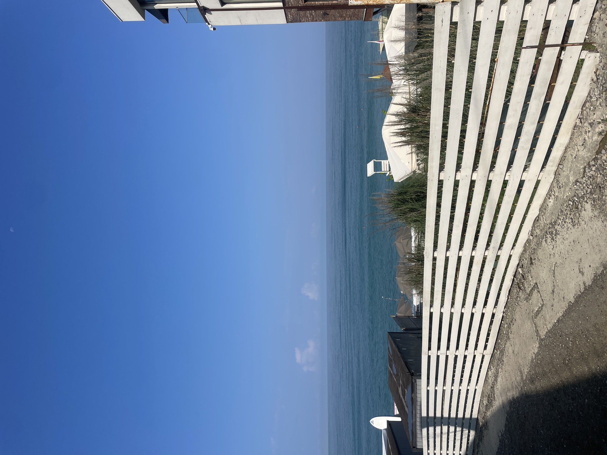 The image depicts a sunlit scene with a clear blue sky. In the foreground, a curved white wooden fence runs across the frame. Beyond the fence, a calm, blue sea stretches to the horizon, meeting the sky with a soft, hazy line. Several white tents or gazebos are visible near the shore, along with some dark, rectangular structures that appear to be storage units or small buildings. A white surfboard stands upright near one of these structures. A modern building with a balcony is partially visible on the right side of the frame. A subtle crescent moon is present in the upper left quadrant of the sky. The overall impression is of a coastal resort area, likely in Sochi, Russia, given the common architectural styles and the sea setting.