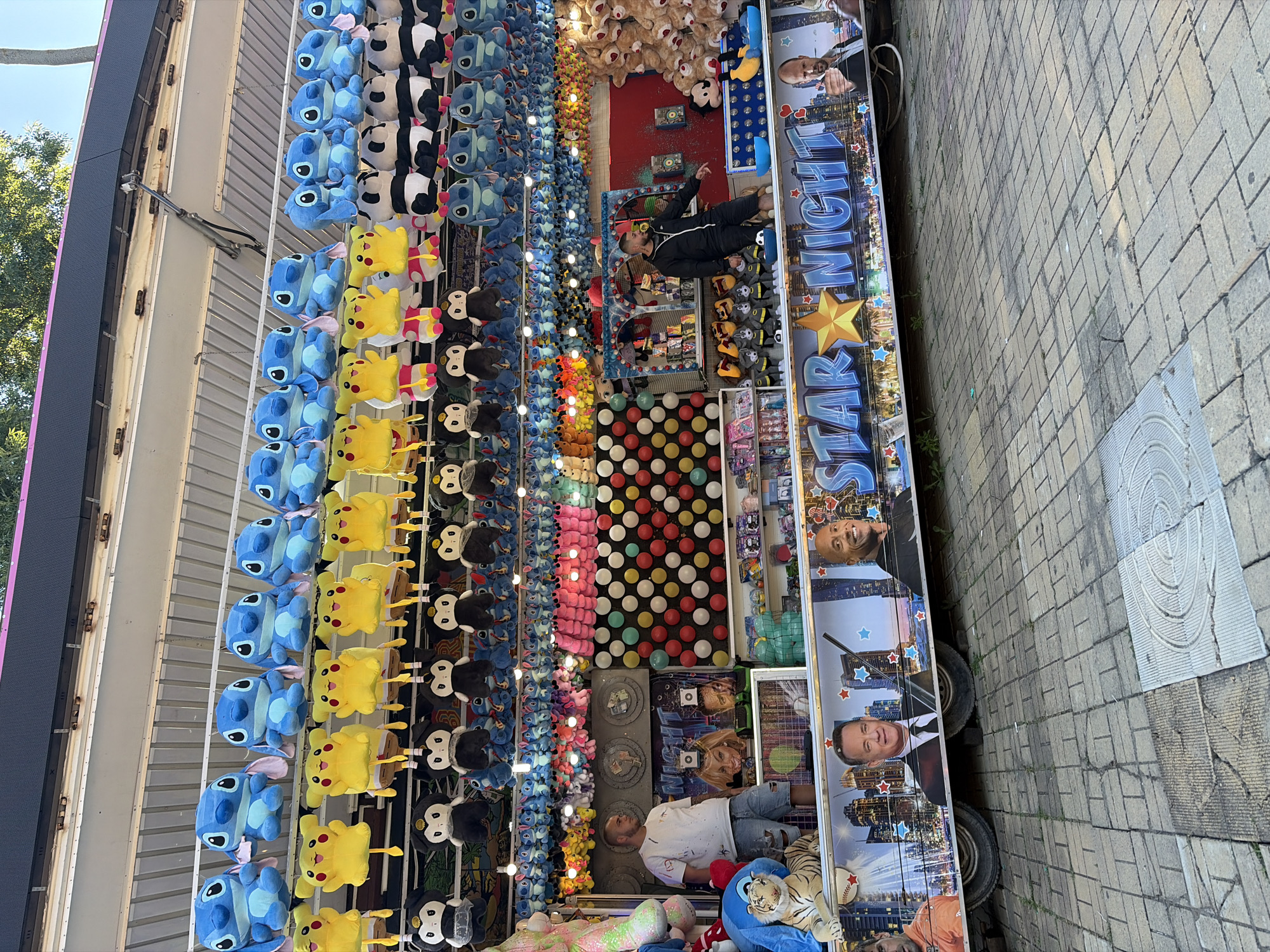 The image shows a carnival game booth with many plush toys for sale, predominantly Stitch characters and Pikachu. The booth features a banner across the top displaying "STAR NIGHT" with images of Tom Hanks and Dwayne "The Rock" Johnson. Below the banner, there are various shelves stocked with more prizes and game elements like colorful balls. Two men are visible; one is a vendor standing to the left, and another is standing to the right near a large pile of plush bears. The ground in front of the booth is paved with grey brickwork. The scene appears to be outdoors during the daytime, with trees visible in the background.