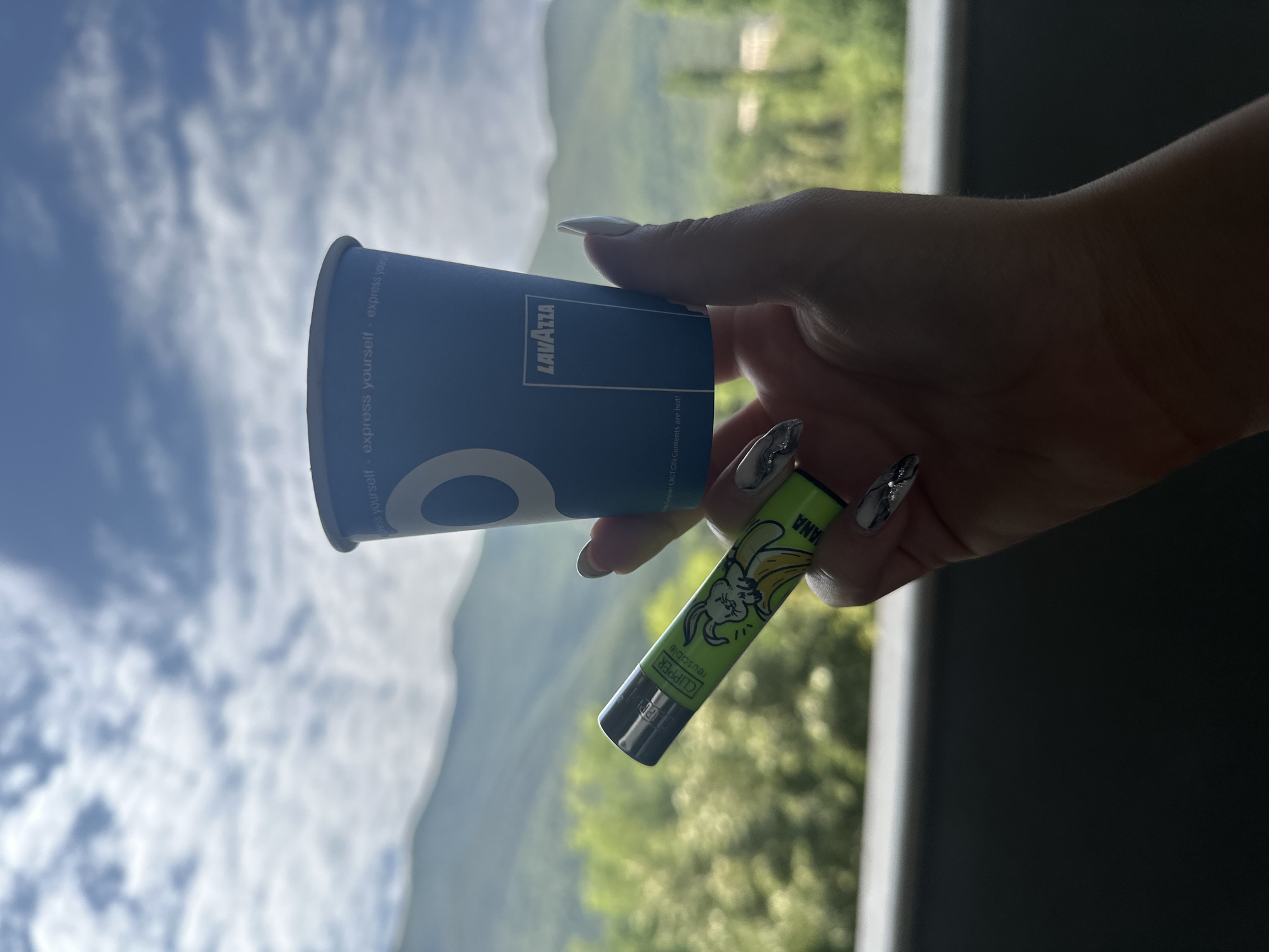 A hand holds a blue Lavazza coffee cup and a green Clipper lighter with a banana and rabbit design against a backdrop of mountains and a cloudy sky. The coffee cup has "express yourself" written in white text and the Lavazza logo. The lighter has "CLIPPER reusable" and "BANANA" written on it, along with a graphic of a rabbit biting a banana. The hand has long, manicured nails with silver glitter. The background suggests an outdoor setting with lush green trees and distant, hazy mountains, possibly indicating a location in or near Vratsa, Bulgaria, given the landscape often associated with the region. The lighting suggests it is daytime, likely afternoon due to the angle of the light.