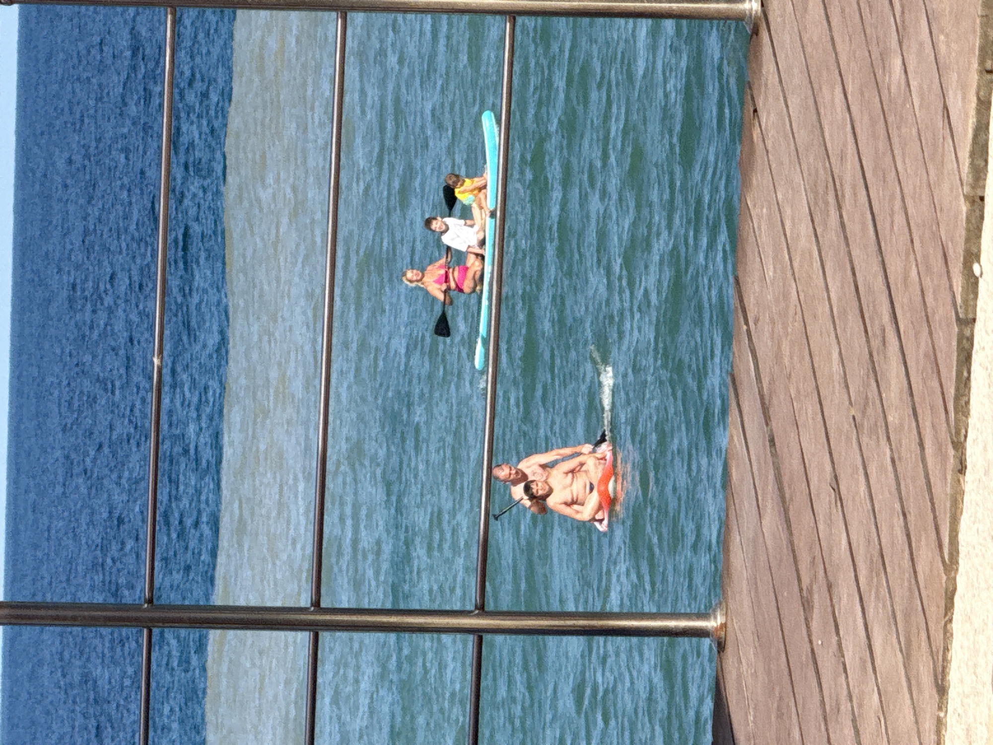 The image depicts a scene at the seaside in Partenit, Ukraine, likely a beach or pier area. In the foreground, a wooden walkway or pier with parallel wooden planks runs diagonally across the frame, leading to the beach and sea. A metal railing, consisting of vertical posts and horizontal bars, partially obstructs the view of the water.
The sea occupies the middle and background of the image, with a distinct line separating two different water colors, possibly due to a river outflow or differing depths. The water is calm, with a slight ripple.
Several people are visible on the water, engaged in recreational activities. On the left side, two shirtless males are on a paddleboard. One man is seated while the other stands behind him, holding a paddle. They appear to be a father and son or an adult and child. On the right side, three individuals, a woman and two children, are on a blue stand-up paddleboard. The woman is wearing a pink bikini and is holding a paddle. The children are seated.
The sky is visible at the very top of the image, appearing bright and clear, suggesting a sunny day. The overall atmosphere is one of leisure and outdoor activity.