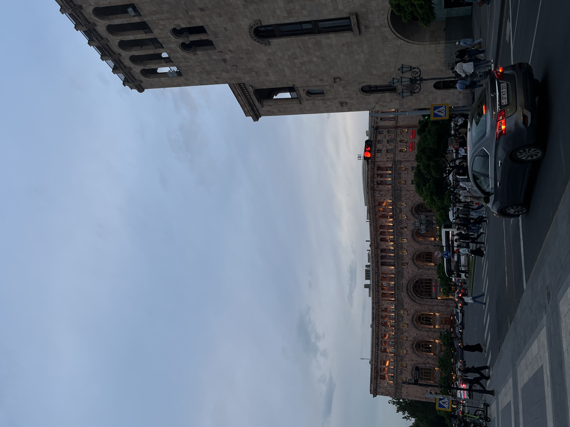 The image shows a street scene in Yerevan, Armenia, at dusk. On the right side of the frame, pedestrians are crossing a street at a crosswalk in front of a large, ornate stone building. A grey sedan is stopped at a red light. The building appears to be constructed of light brown stone, with multiple floors and arched windows. Streetlights are visible. On the upper left, there's another stone building with a similar architectural style. The sky is overcast. The car's license plate reads "37 KO 111".