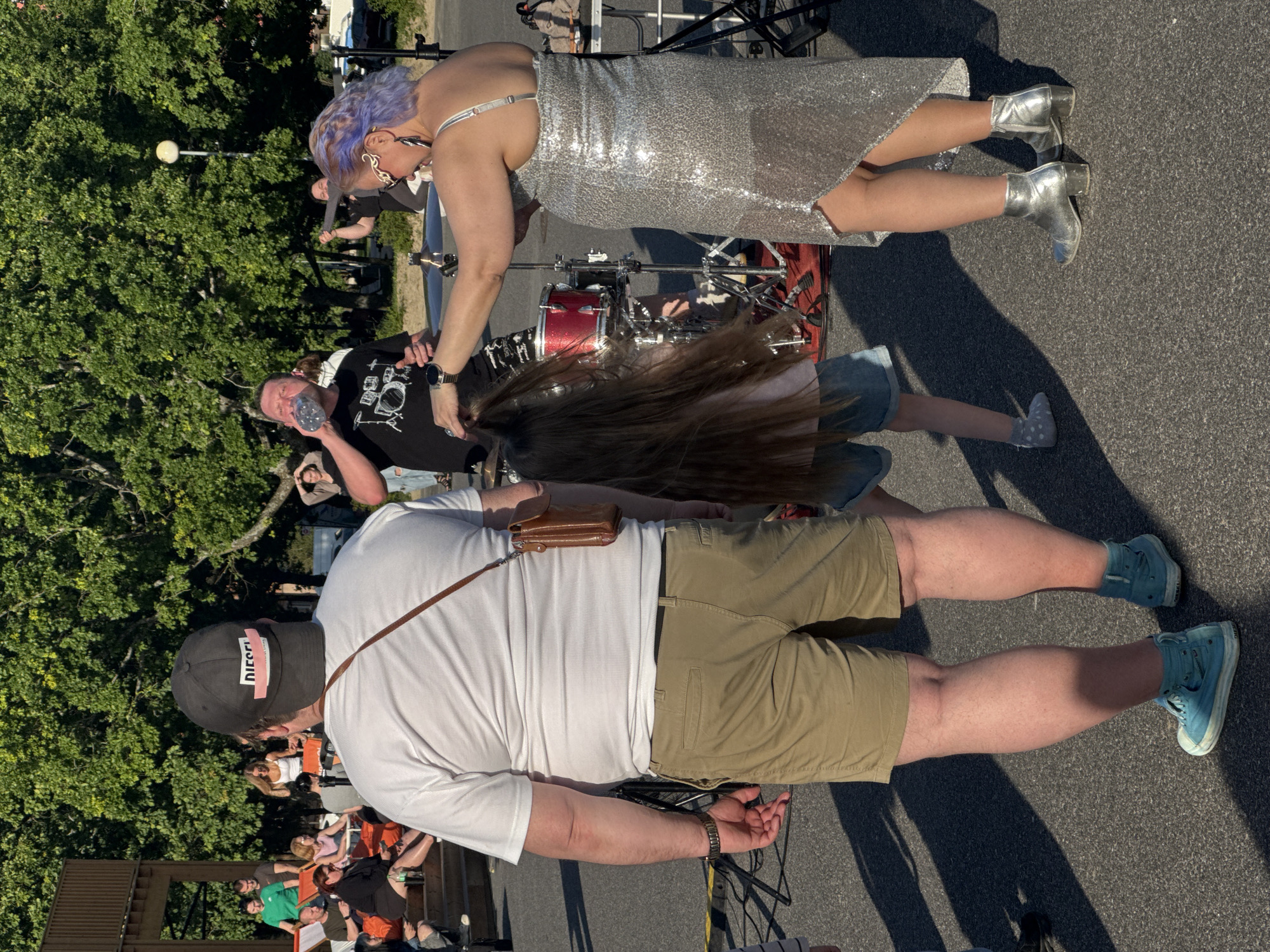 In Tallinn, Estonia, at an outdoor event, a person wearing a white t-shirt, khaki shorts, and light blue high-top sneakers stands facing away from the camera, with a brown leather pouch attached to a strap across their chest. To their right, a young girl with long brown hair is partially visible. In the foreground, a woman with purple hair styled in an updo, wearing a shimmering silver dress and silver ankle boots, is interacting with the girl, appearing to brush or style her hair. Behind them, a person in a black t-shirt with a drum set graphic is drinking from a clear plastic bottle. A drum set is set up on a red rug. Other individuals are visible in the background, some seated on bleachers or standing amidst trees. The ground is paved, and the scene is illuminated by sunlight, casting shadows.