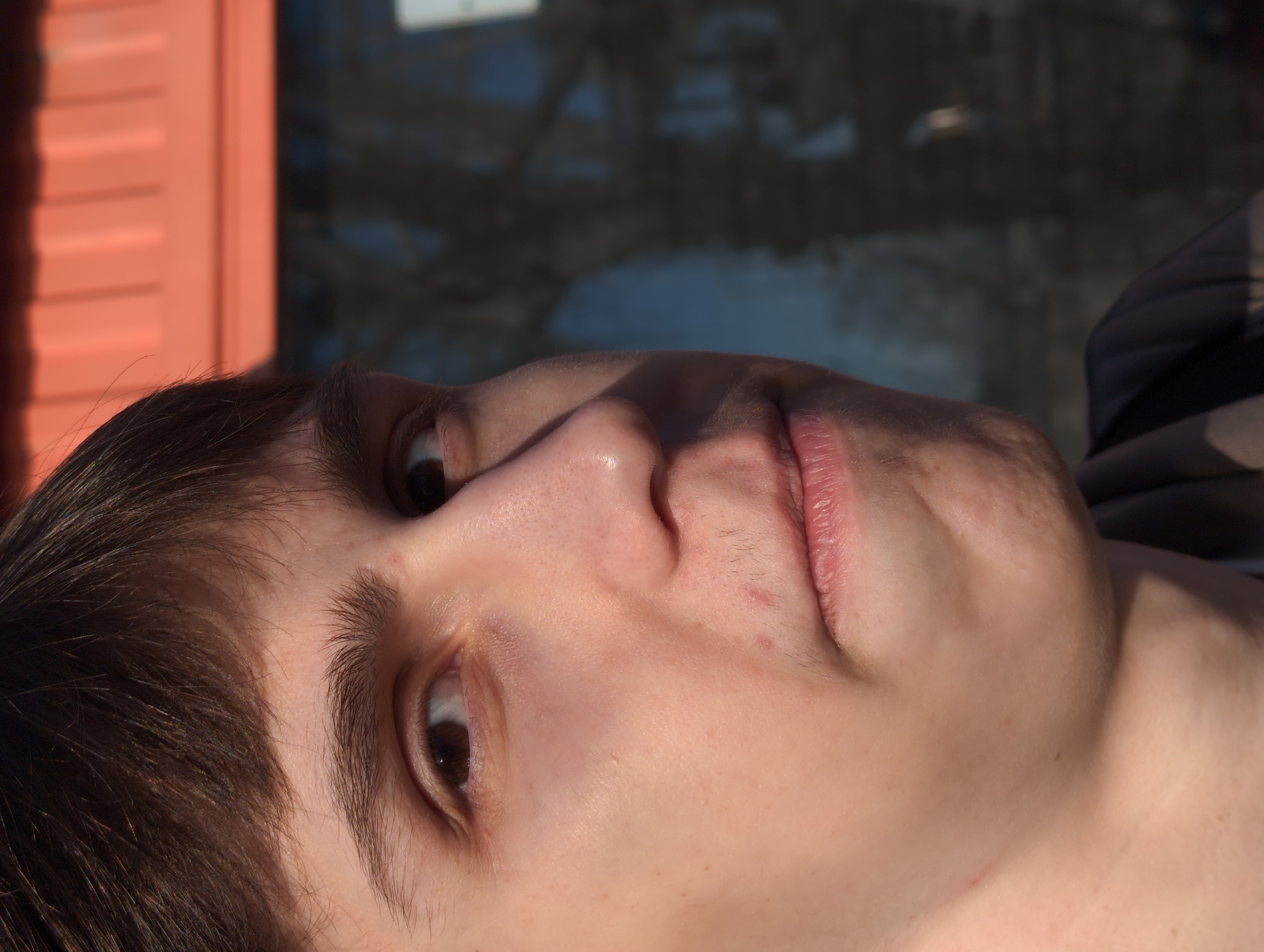A close-up, upward-angled shot focuses on a young person's face. The person has brown hair and brown eyes. Skin is light-toned. Hints of peach fuzz can be seen above the lips. The background is blurred, with hints of an orange or red-colored structure with vertical panels on the left and a darker, indistinct structure behind the person. No other people, objects, or actions are visible. The location of the city of Izhevsk, Russia is not visible in this image.