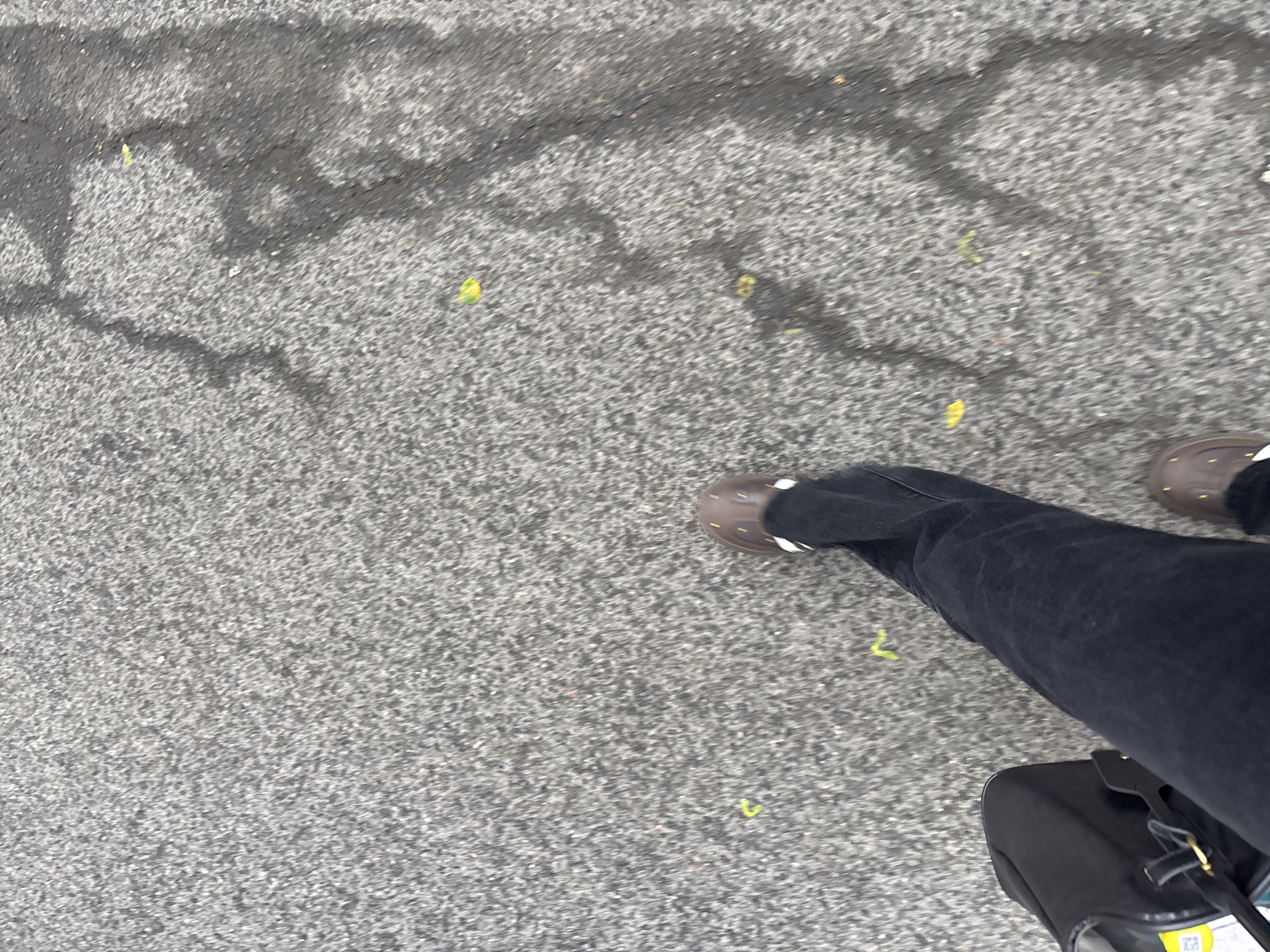 A ground-level shot shows a section of cracked asphalt pavement in what appears to be an urban area. Scattered across the asphalt are several small, yellow, linear objects, possibly small twigs or debris. A person's legs and feet are visible in the bottom right portion of the image, clothed in black jeans and brown shoes. A black bag is positioned next to the leg. The location is unknown, but could be Russia.
