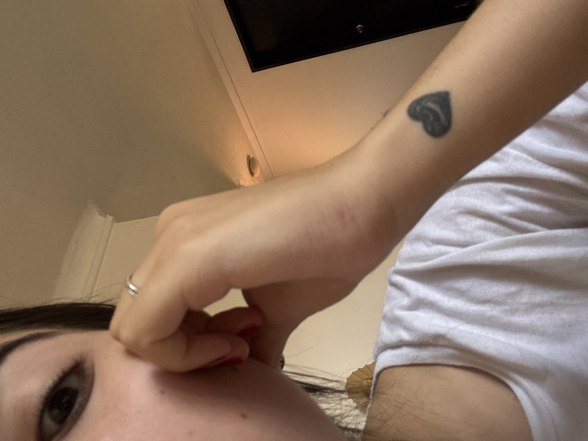 A low-angle shot of a person's face, forearm, and chest. The person has dark hair and is wearing a white t-shirt. Their fingers are touching their face near their nose, and they are wearing a ring on one finger. On the forearm, there is a black heart tattoo. In the background, there is a ceiling and an obscured black object. It is not possible to infer the location as Oaxaca City, Mexico, from the given image.