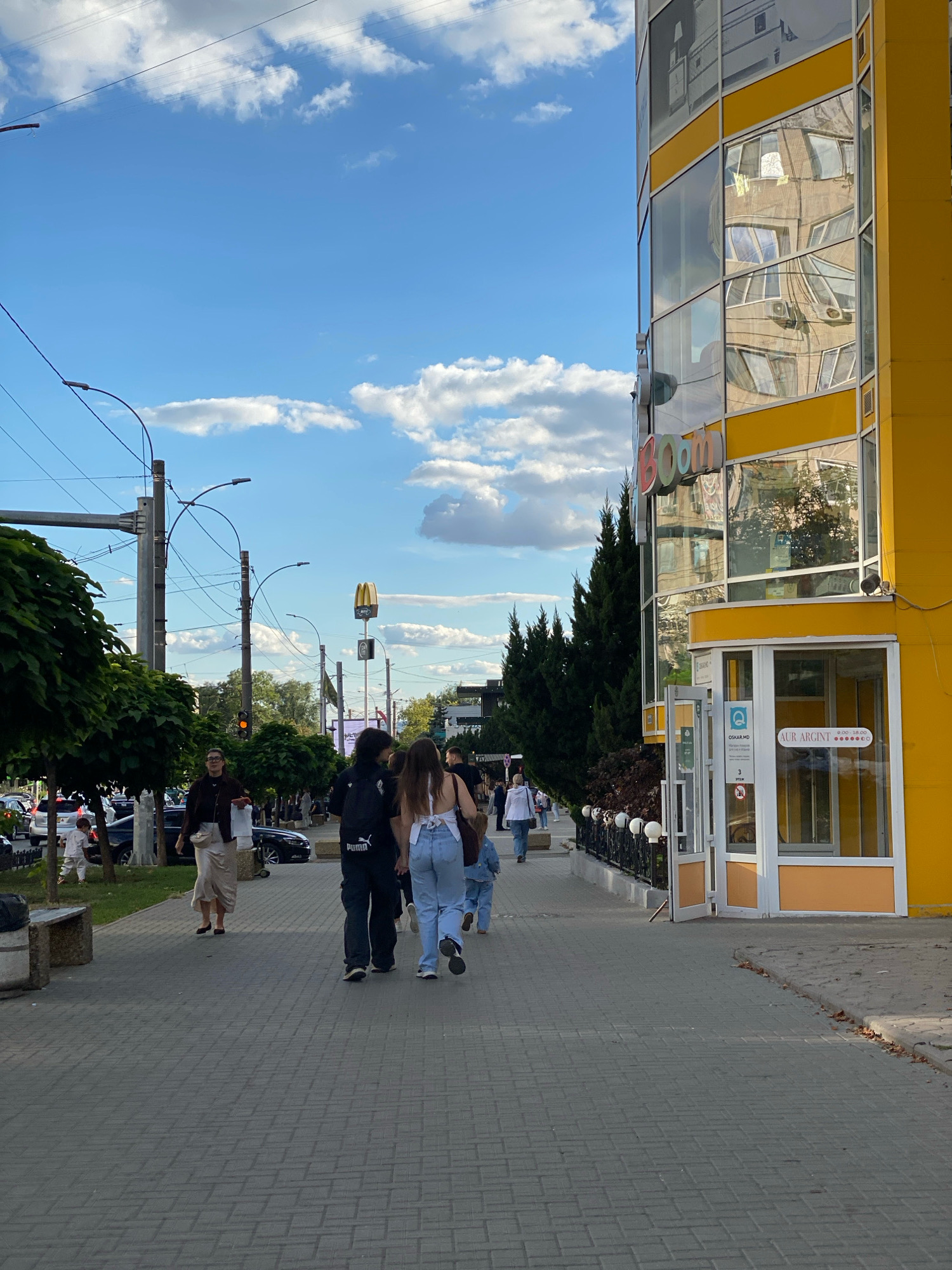 This image depicts a street scene in Chișinău, Moldova, likely during daytime. A bright yellow building with extensive glass windows dominates the right side of the frame, reflecting the surrounding environment and sky. A sign with colorful letters, partially visible, reads "Boom". Pedestrians are walking along a paved sidewalk. In the background, a McDonald's sign is visible, suggesting the presence of a fast-food restaurant. Traffic can be seen on the street to the left, with several cars present. Trees line the sidewalk, and streetlights and utility poles are also visible. The sky is blue with scattered white clouds.