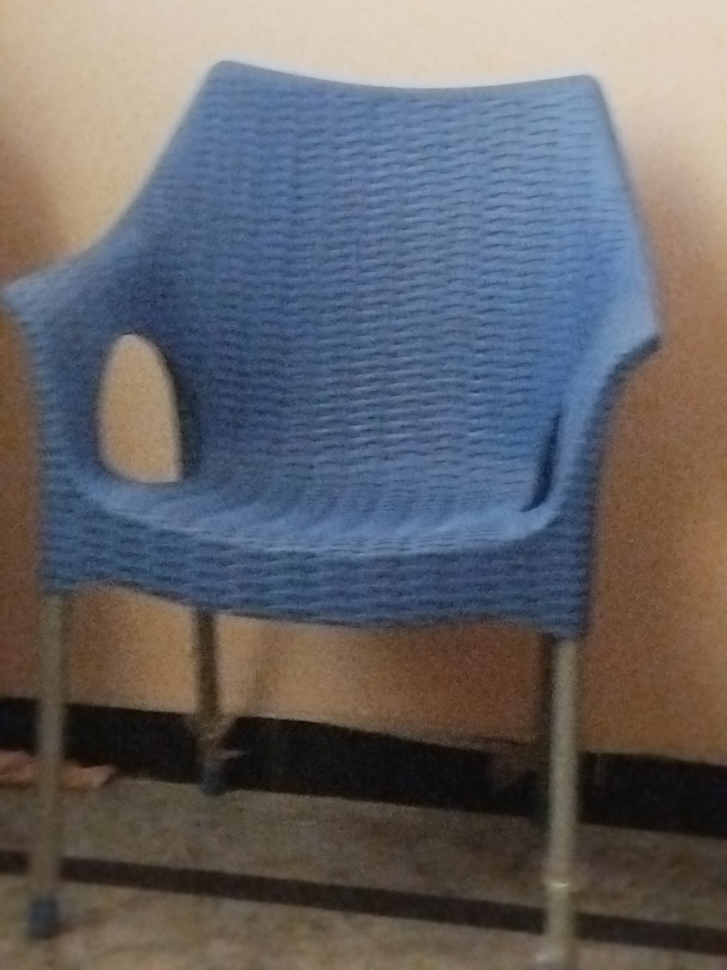 A blue plastic chair with a woven texture sits on a tiled floor against a beige wall. The chair has a cutout on each side, allowing for armrests. The legs of the chair are silver. The floor is tiled with a pattern of light and dark grey squares. A black line runs across the floor behind the chair. The lighting suggests an indoor environment, possibly a room. The location is identified as Lassan, Pakistan.