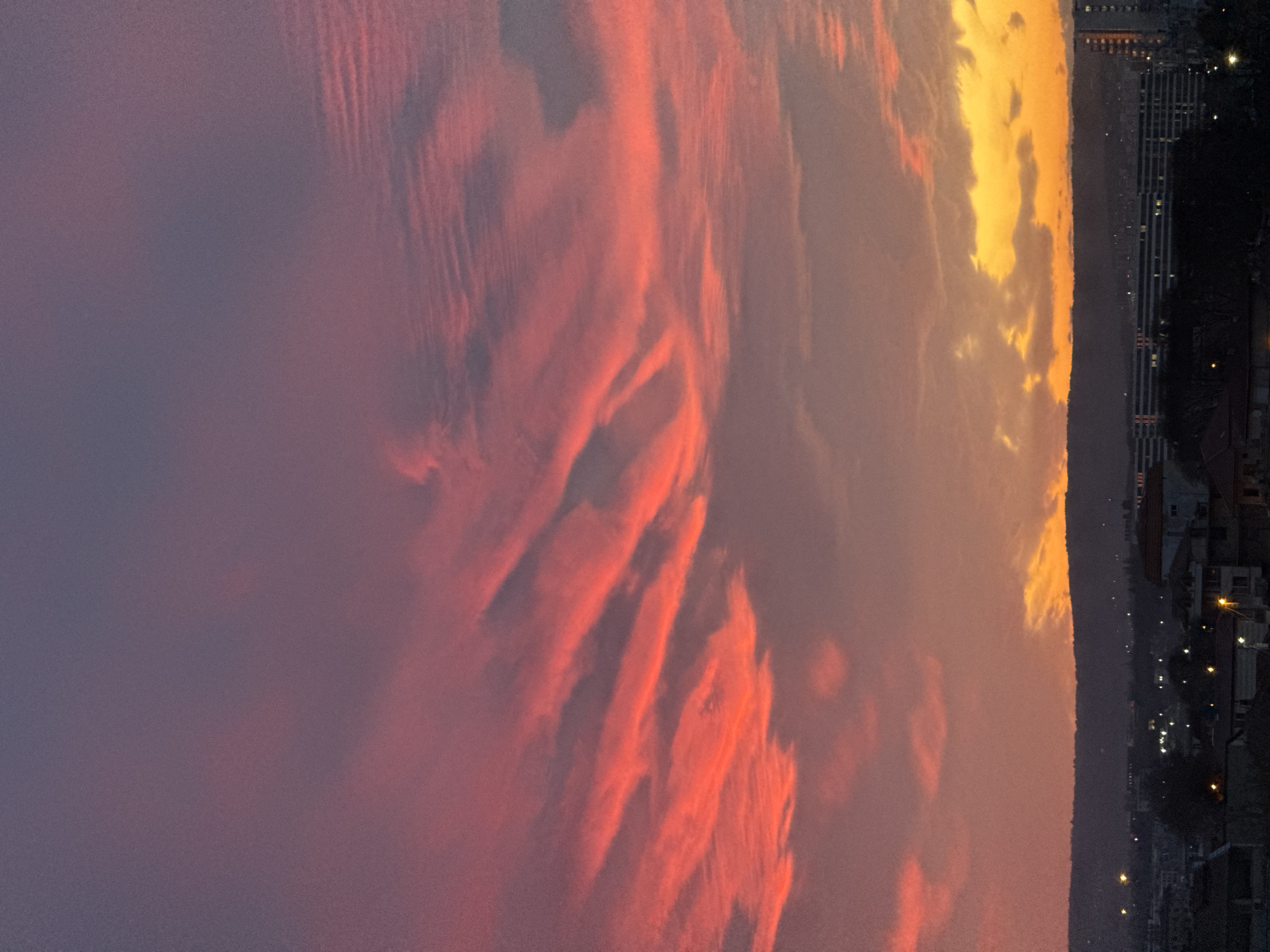 The image depicts a twilight cityscape under a dramatic, colorful sky. The sky transitions from a deep purple on the left to orange and yellow hues towards the right. Altocumulus clouds dominate the view, catching the light of the setting sun in various shades of red.
At the bottom of the image, a dark strip represents the cityscape of Varna, Bulgaria. The dark silhouette of the land shows buildings of varying heights, with scattered lights indicating individual windows and street illumination. The lights appear more densely populated in certain areas.
The absence of recognizable human figures or active events focuses the scene on the interplay of natural light and urban setting. The orientation of the image is horizontal. The sky covers the vast majority of the view and casts an ambient glow over the buildings below.