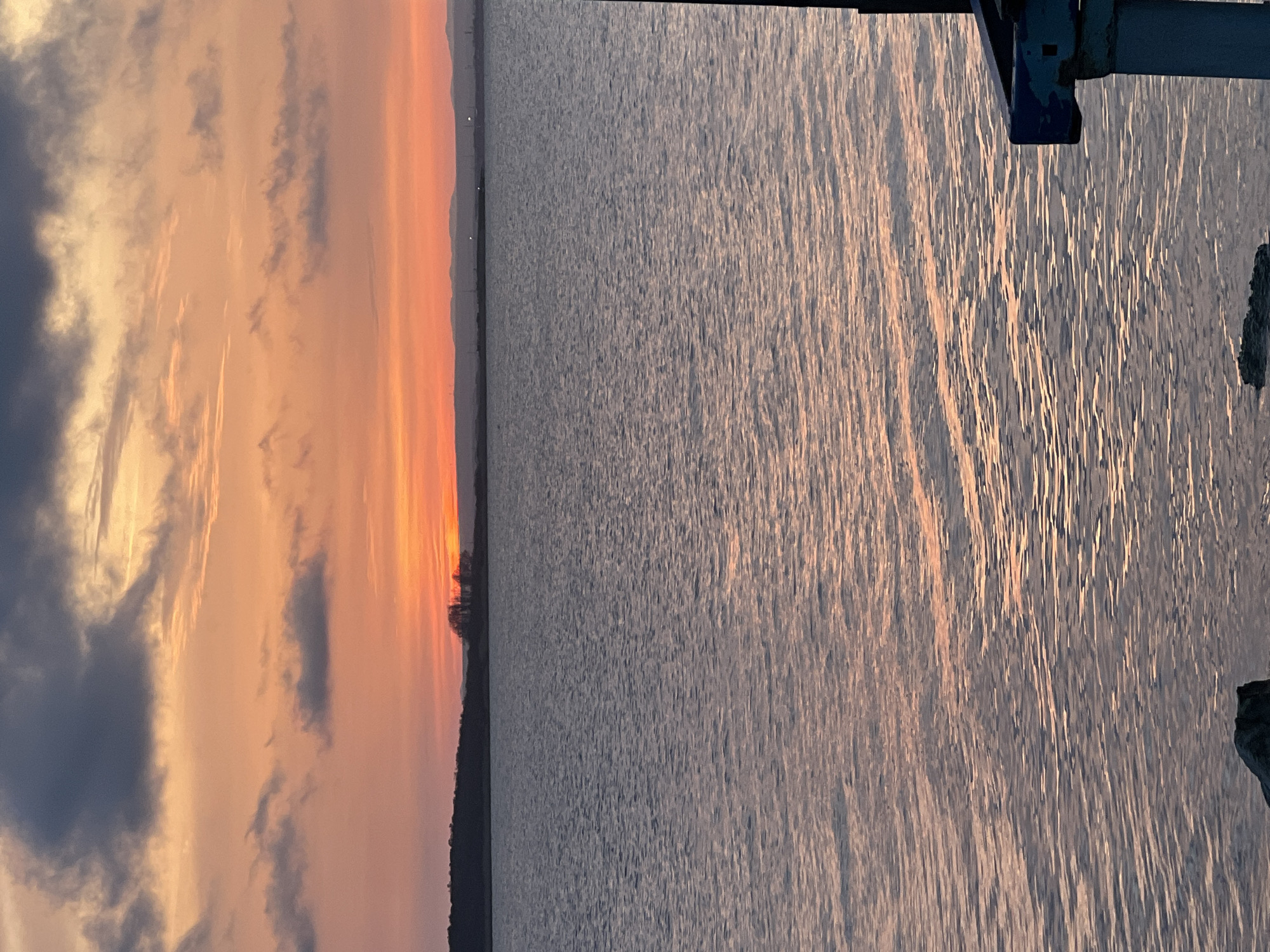 The image captures a vibrant sunrise or sunset over a vast body of water, likely the Black Sea near Burgas, Bulgaria. The sky, occupying the upper half of the frame, is awash with warm hues of orange, pink, and golden yellow, particularly near the horizon where the sun's glow is most intense. Several clouds, ranging from soft, wispy formations to darker, denser masses on the left, are scattered across the sky, adding texture and drama to the colorful display.
Below the horizon, the expansive body of water reflects the sky's light, creating shimmering, elongated streaks of orange and yellow across its surface. The water has a textured appearance, indicating gentle ripples or small waves catching the sun's rays. A thin strip of distant land or coastline is visible along the horizon, silhouetted in dark tones, with a distinct cluster of trees rising above it on the left side of this distant landmass.
On the far right side of the frame, a dark, angular man-made structure, possibly part of a pier, dock, or boat, extends into the scene. This structure provides a foreground element, hinting at human activity or infrastructure in the area. The overall atmosphere is serene and picturesque, emphasizing the beauty of the "golden hour" light over the water. No visible text is present in the image.