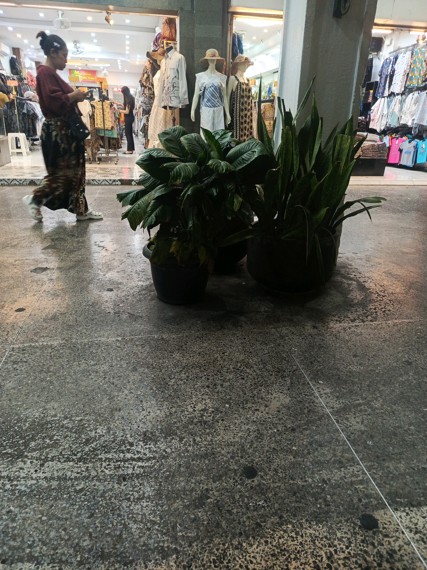In Yogyakarta, Indonesia, a woman with her hair in a bun and wearing a maroon top and a patterned skirt walks from left to right in front of a row of shops. She is looking down at her phone. In front of the shops, two potted plants are visible. The shops display clothing on racks and mannequins, including patterned shirts, dresses, and t-shirts. The floor is a rough concrete with a light grey speckled pattern. The lighting suggests it is evening or indoors.