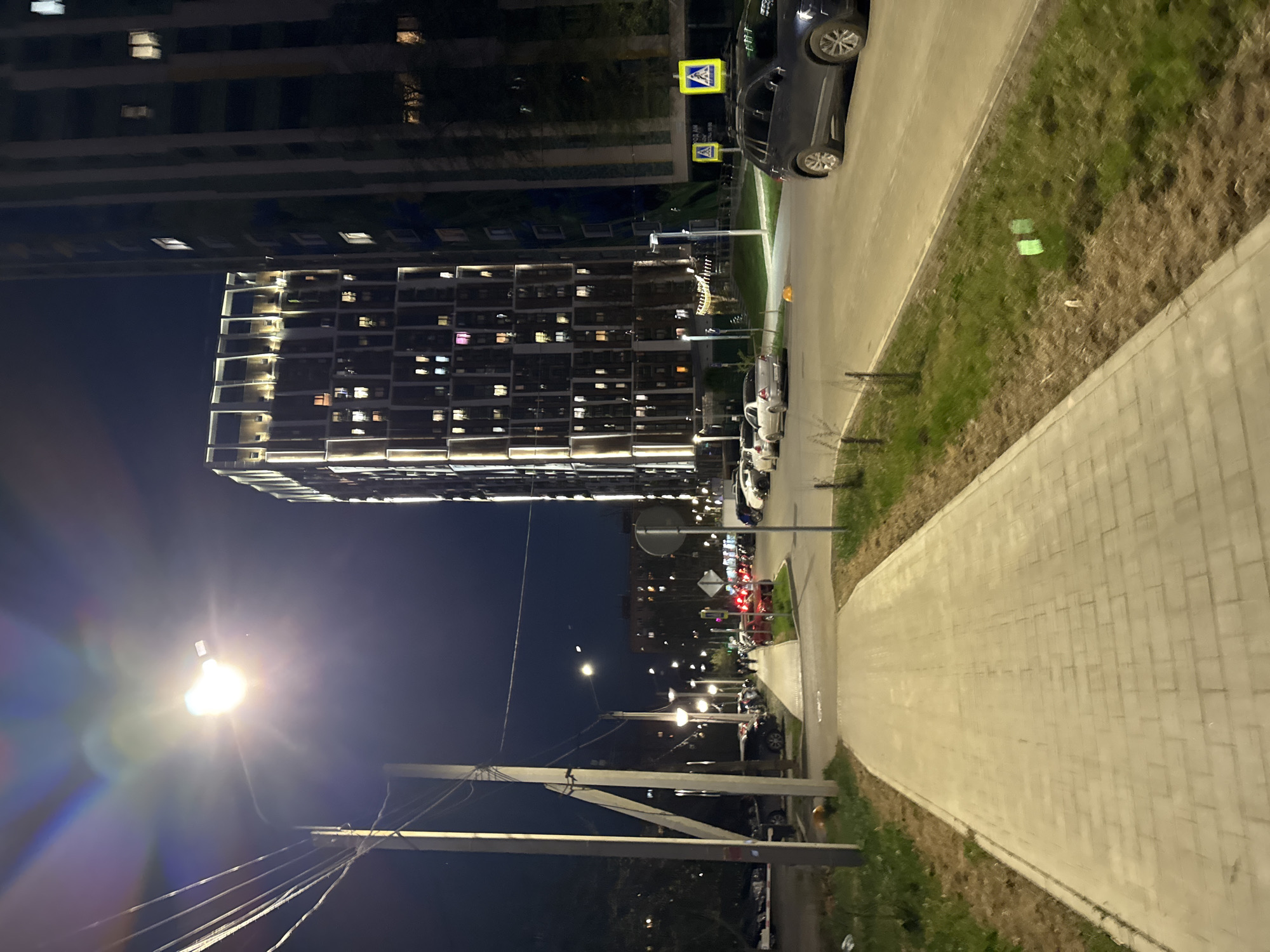 The image depicts a nighttime street scene in Izhevsk, Russia. A large, modern apartment building with illuminated windows is visible in the background. A row of parked cars lines the street. Streetlights cast a bright glow. A wide, paved sidewalk runs along the right side of the image. Utility poles and wires are visible in the lower part of the frame. Vegetation is present along the sidewalk and roadside. Pedestrian crossing signs are visible near the parked cars.