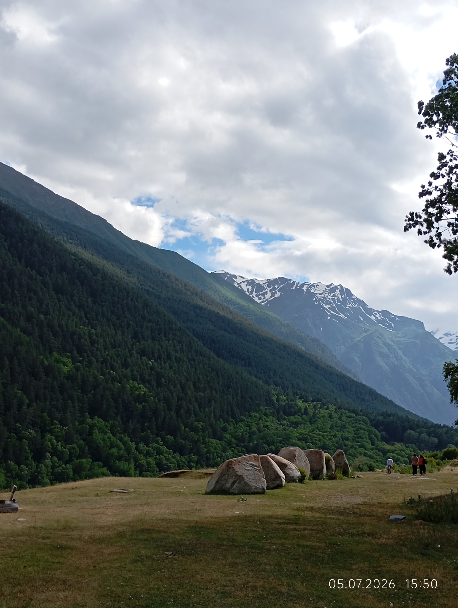 The image depicts a mountainous landscape with a dense forest covering a steep slope on the left. Snow-capped peaks are visible in the background, indicating a high-altitude environment. In the foreground, several large, rounded rocks are arranged in a semi-circle on a grassy field. Three people are walking in the distance towards the right side of the frame. The sky is partially cloudy. A date and time stamp, "05.07.2026 15:50," is present in the lower right corner. The scene is consistent with a location in or around Neutrino, Russia, given the characteristic mountain ranges and vegetation often found in such areas.