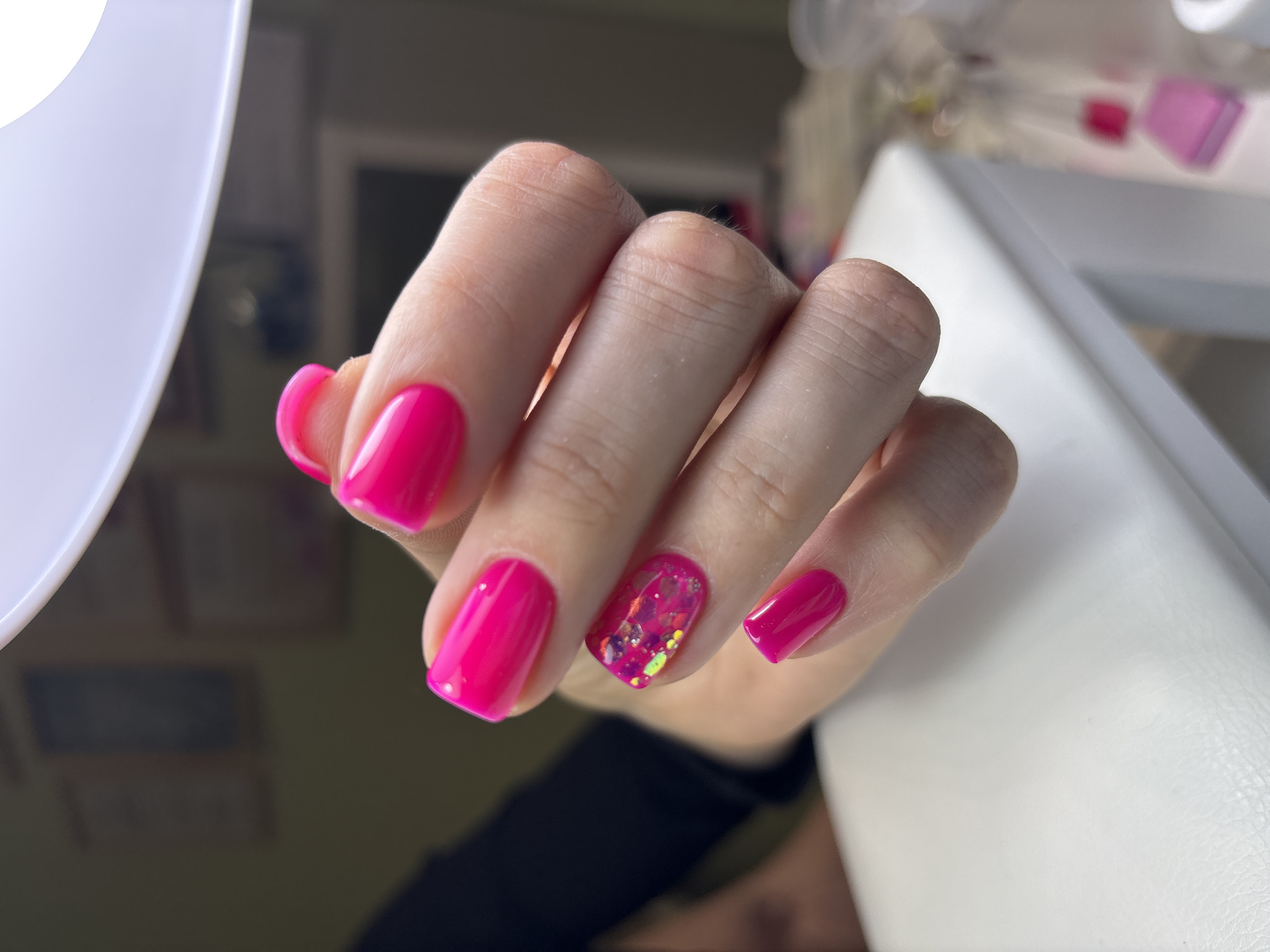A close-up shot features a hand with freshly manicured nails, predominantly painted in a bright pink color. One nail has a glitter design in a similar pink hue. The hand is resting on a white surface, possibly a table or cushion. In the background, a blurred setting suggests a room with various objects and furniture. No specific location or activity is discernible. There is no indication of the city of Ферма, Russia.