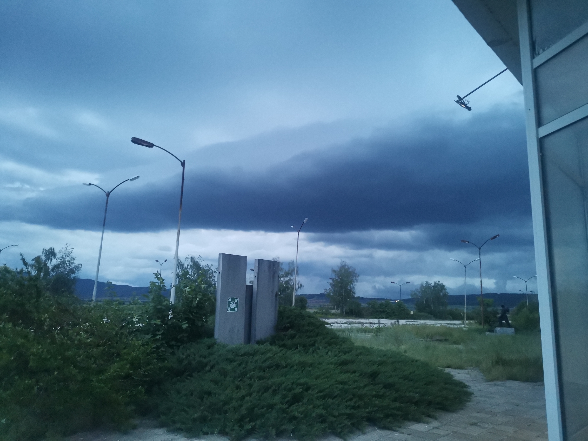 The image presents a desolate outdoor scene in Zhivkovo, Bulgaria, under a dramatically dark and cloudy sky. Heavy, blue-grey clouds dominate the upper portion of the frame, suggesting an impending storm or very overcast conditions, likely at dusk or dawn.
The foreground is characterized by dense, overgrown dark green bushes and wild grass, indicating a neglected area. A paved path or concrete slab surface is visible on the lower right, leading towards the partial view of a building on the far right, which features a white-framed structure and a paneled wall. An angled metal fixture extends from this building.
Throughout the midground, several tall, unlit streetlights stand amidst more wild greenery. Two prominent, weathered concrete slab-like structures, possibly part of a monument or an old sign, are central in the midground. One of these structures displays a green and white symbol, resembling an emergency exit sign with a white figure walking through a door with an arrow. Further back, partially obscured by vegetation, a dark, human-like statue on a pedestal can be seen.
In the distance, dark mountain ranges are visible beneath the brooding sky. The scene is devoid of any discernible human or animal activity, conveying a sense of quiet abandonment and an eerie stillness, intensified by the stormy weather.