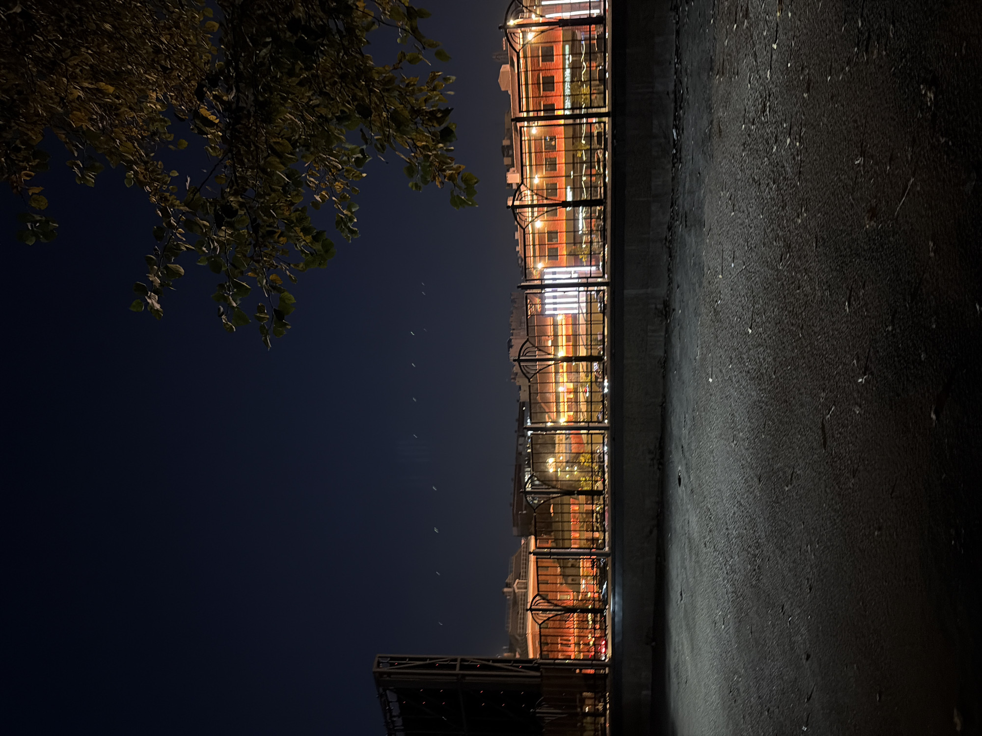 The image shows a night scene in Moscow, Russia. In the foreground, there is a dark asphalt surface with some scattered debris. A concrete barrier runs along the middle of the image, separating the foreground from a fenced area behind it. Beyond the fence, a multi-story building with red brick facade is illuminated by warm orange and white lights. The building appears to be a commercial establishment, possibly shops or offices, with visible windows and architectural details. The fence itself is made of metal with decorative patterns. In the sky, faint white streaks suggest moving lights, possibly from aircraft or streetlights. To the right of the frame, a tree with dark leaves overhangs the scene. The overall atmosphere is urban and nocturnal.