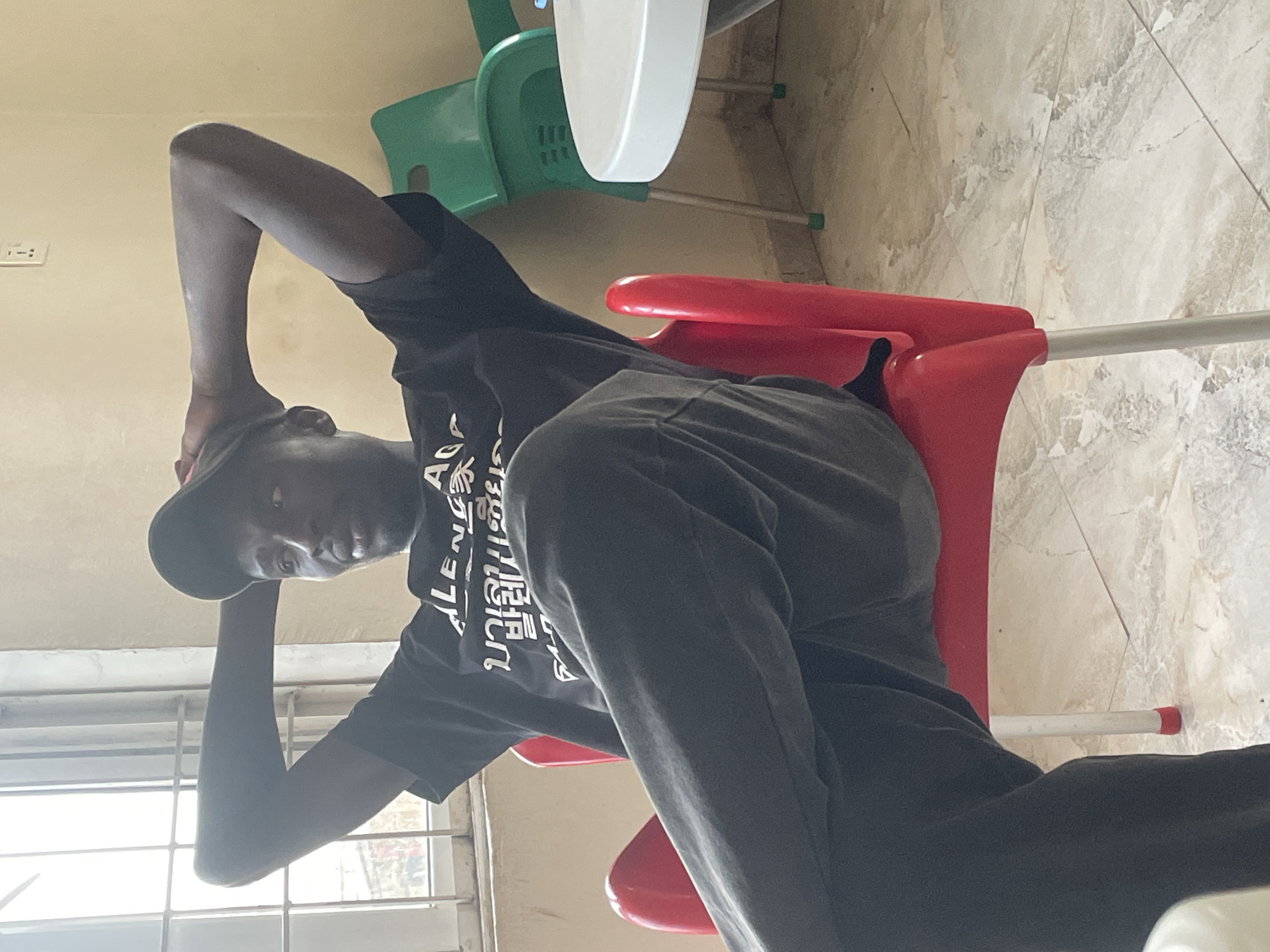 A young man is seated in a red plastic chair. He is wearing a black T-shirt with white lettering and dark sweatpants. He has a black cap and is positioned with his hands behind his head, leaning back. The background shows a window with metal bars, a light-colored wall, and a tiled floor. In the background, there is also a green plastic chair with a white plastic table attached. The image appears to be taken indoors. No definitive indication is present that would lead to determining the location is specifically in Jos, Nigeria.