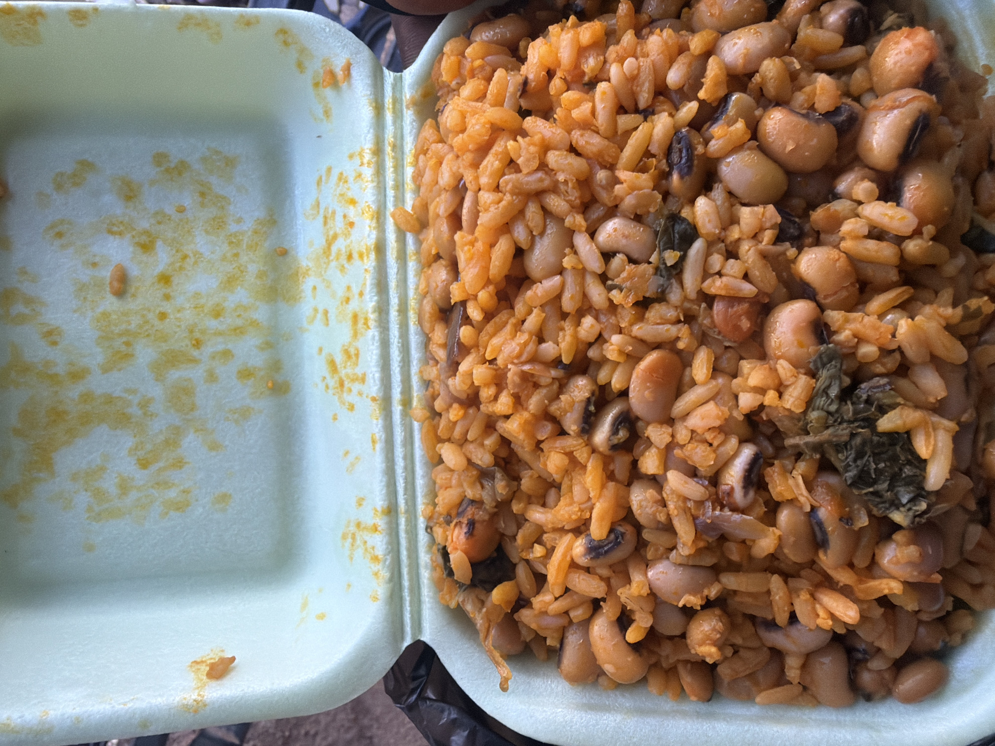 A close-up, overhead shot shows a light green styrofoam takeout container filled with a vibrant orange dish of rice and beans. The lid of the container is partially open, revealing the appetizing meal within. The rice grains are small and slightly separated, coated in a rich orange sauce, and mixed with an abundance of light brown beans, many of which have a distinctive black spot. Scattered throughout the dish are small pieces of dark green leafy vegetables and some darker, possibly dried, ingredients. The interior of the container's lid is stained with remnants of the orange sauce, suggesting the dish was recently served. The background is blurred and out of focus, but appears to be an outdoor setting with muted earthy tones. The image is sharp and well-lit, highlighting the texture and color of the food. This dish, likely a local specialty, is presented in a common takeout format, suitable for a quick meal.