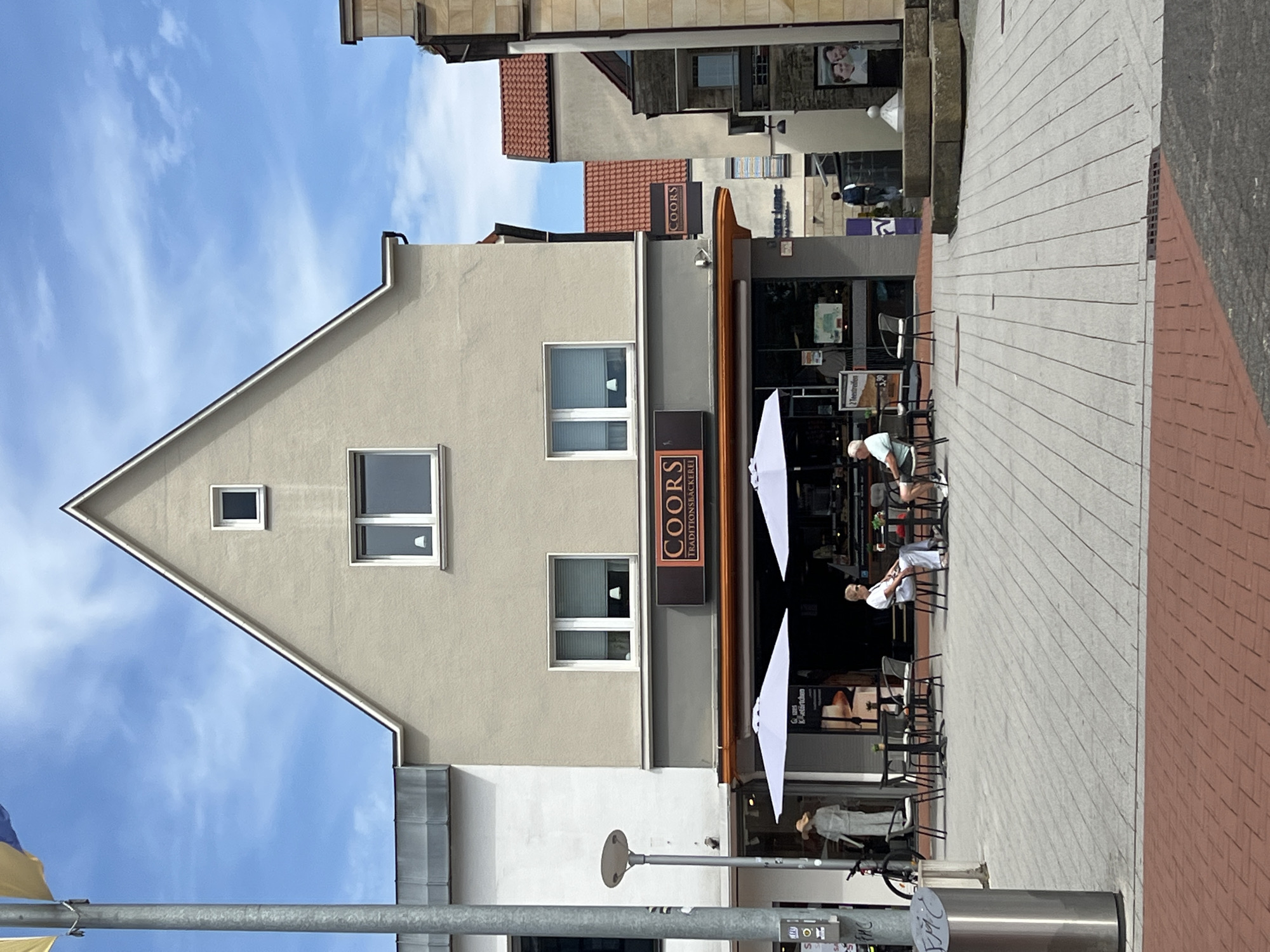 In the city of Unknown, Unknown, a two-story building with a beige facade and a gable roof stands on a cobblestone plaza. The building features multiple windows, with two on the upper level and two on the lower level, each with white blinds. A prominent sign above the ground floor entrance reads "COORS TRADITIONSBÄCKEREI" in white lettering on a dark brown background. The ground floor houses a bakery with an outdoor seating area. Two white umbrellas provide shade for tables and chairs where two individuals, one male and one female, are seated. The male, wearing a white t-shirt and light-colored pants, is looking to his right. The female, also in a white shirt and light pants, is facing towards the bakery. A mannequin dressed in a straw hat and a light-colored outfit is visible through the shop window on the left. Adjacent to the bakery, a grey trash receptacle and a bicycle are present. The plaza is paved with light grey and red bricks, and a flagpole with two yellow and blue flags stands to the left of the building. In the background, other buildings with red tile roofs are visible, along with a public fountain and a poster featuring two people. A person wearing a backpack is walking away from the camera in the distance. The sky above is blue with scattered white clouds.