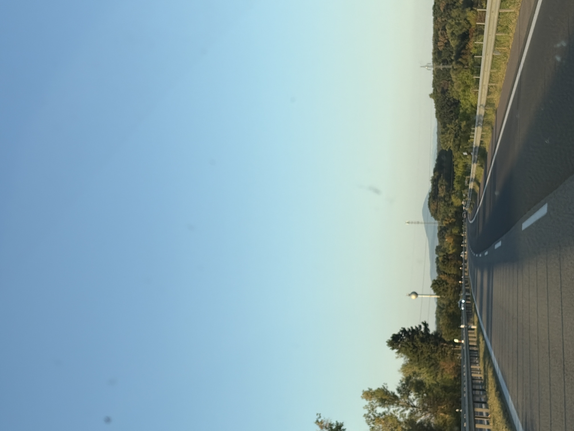 The image depicts a perspective view from a vehicle traveling on a highway. The highway has multiple lanes separated by white lines, and a metal guardrail runs along the side. The surrounding environment consists of trees and vegetation, with a clear blue sky overhead. In the distance, a water tower and a transmission tower are visible, suggesting a semi-rural or suburban setting. The lighting indicates it is daytime, likely late afternoon or early morning due to the soft sunlight. No people are visible in the image, and the focus is on the road and landscape. The scene is consistent with a location on a highway near the town of Veľké Zálužie, Slovakia, characterized by its infrastructure and natural surroundings.