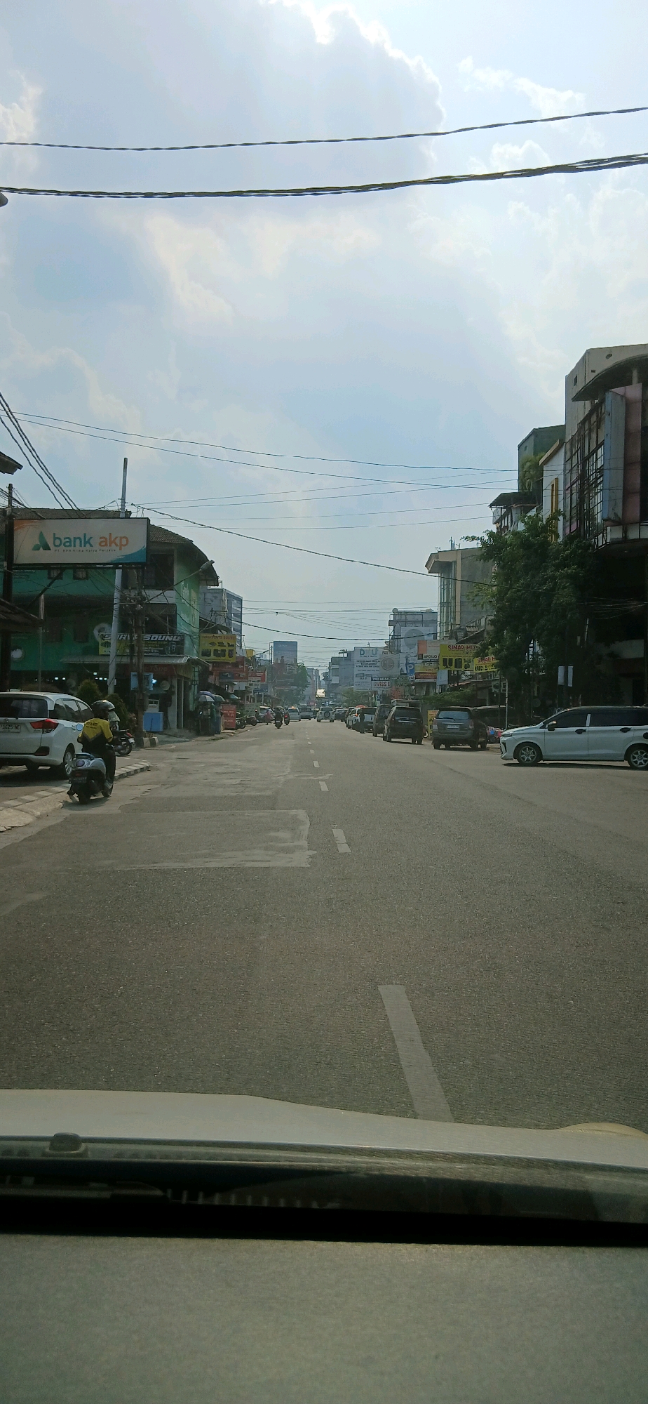 This is a vertical, medium-shot, eye-level, full-color photograph taken from the perspective of someone in a vehicle, looking down a paved road in Samarinda, Indonesia. The road is lined with buildings on both sides, and there is moderate traffic. The sky is cloudy and bright, indicating it is daytime.
On the left side of the road, there are several buildings, including one with a prominent sign that reads "bank akp." Below this sign, another sign appears to say "STAVE SOUND." There are parked cars and motorcycles in front of these buildings. A person wearing a yellow jacket is sitting on a motorcycle near the curb.
On the right side of the road, more buildings are visible, some with multiple stories. Trees are interspersed among the buildings. There are several cars driving on the road, some parked. A white sedan is parked on the right shoulder of the road.
The road itself has white dashed lines marking lanes. The asphalt appears to be in fair condition with some patches. Overhead power lines are visible crisscrossing the sky. The overall impression is of a busy urban street during the day.