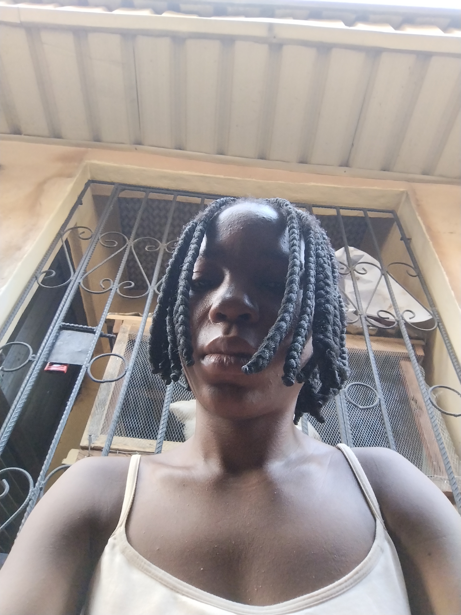 A low-angle, medium shot captures a person from the shoulders up, facing the camera. They have dark skin and dark hair styled in numerous braided pigtails that fall forward over their shoulders and partially obscure their face. The person is wearing a light-colored camisole. Behind them, a barred window is visible, with decorative metalwork. The wall surrounding the window is textured and appears to be painted a light brown or tan color. Above the window, a white corrugated metal roof is visible, casting shadows. The overall impression is of an outdoor setting, possibly a residential area, with a focus on the individual and the architectural elements behind them. The scene is set in Isiehor, Nigeria.