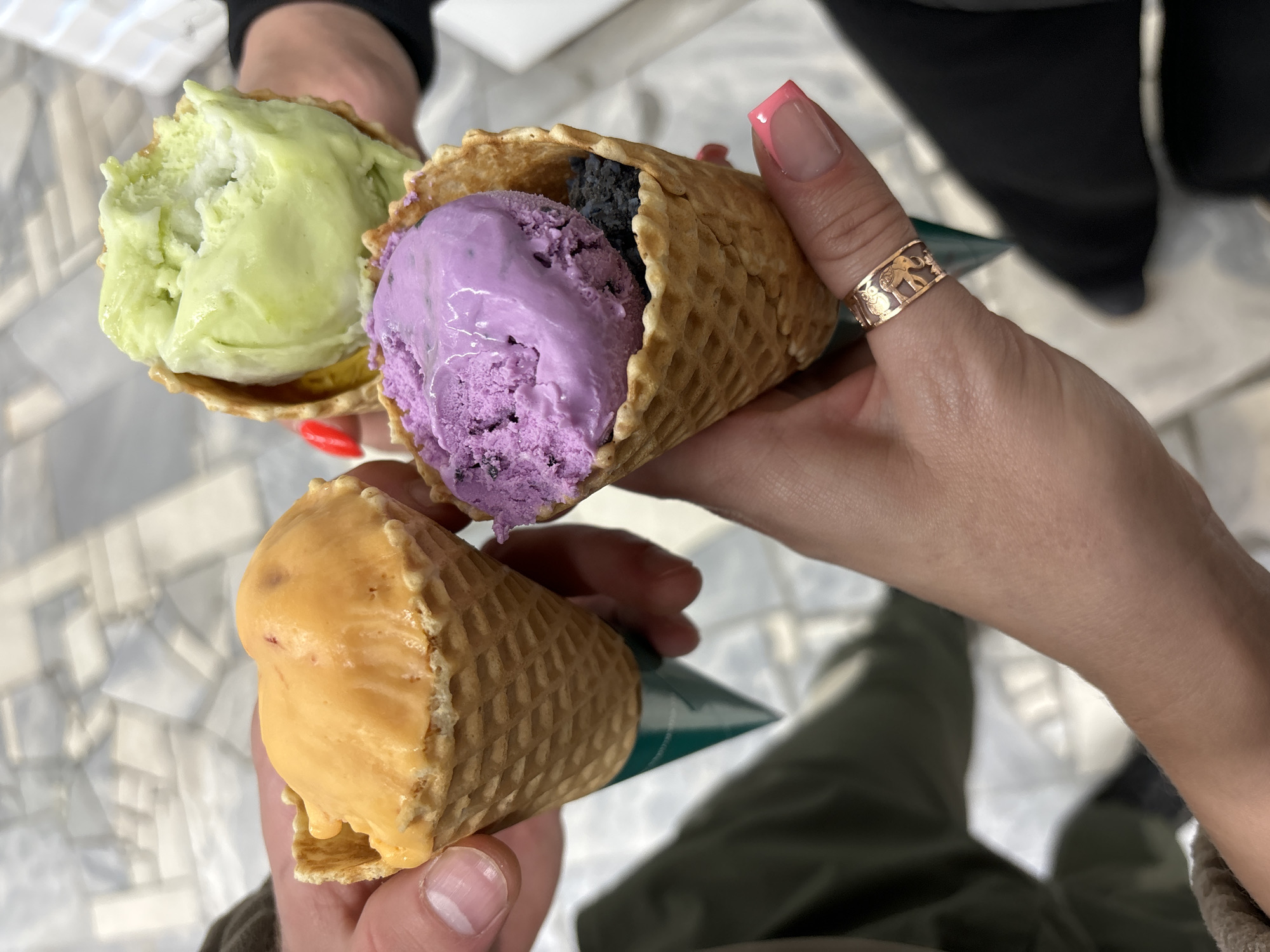 A close-up, high-angle shot captures three hands holding waffle ice cream cones indoors in Chaikovsky, Russia. Three distinct ice cream flavors are visible: a bright lime green scoop, a vibrant purple scoop accompanied by a smaller, very dark (possibly black or dark grey) scoop, and an orange-yellow scoop. The cones are classic waffle cones, each with a dark green paper wrap at the bottom. One person's hand, positioned centrally, displays manicured nails with pink tips and a golden ring on the thumb, featuring intricate elephant designs. The background reveals a light-colored, tiled floor with a geometric pattern, suggesting an indoor setting like a cafe or ice cream parlor. The image depicts a casual moment of individuals enjoying ice cream treats. No specific time of day or weather is discernible, and there is no visible text.
