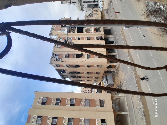 A damaged urban landscape with buildings showing signs of war damage like bullet holes and structural decay. A person is seen in the street. The image is taken from an elevated position, possibly a high-rise building, looking down onto the scene through a rusty metal structure. The sky is overcast.