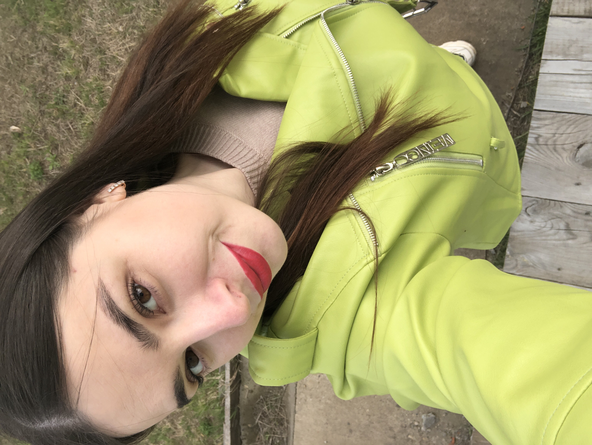 A selfie-style portrait, captured outdoors in Noviy, Russia, features a young woman looking directly at the camera with a subtle, engaging smile. Her head is tilted, and her long, dark brown hair frames her face and cascades over her shoulders. She wears a striking, bright lime-green or chartreuse leather-like jacket with numerous prominent silver zippers, and a light beige or brown ribbed knit sweater underneath.
Her facial features include full eyebrows, dark brown eyes with visible eyelashes, and bright red lipstick. A small, sparkling stud earring is visible in her right ear. One of the jacket's zipper pulls clearly displays the text "VEINCON". The background is softly blurred but reveals an outdoor setting, with patches of green grass, brown earth, and wooden planks or a pathway. A white shoe is partially visible in the upper right. The bright, even lighting suggests it is daytime. The image conveys a casual and confident demeanor.