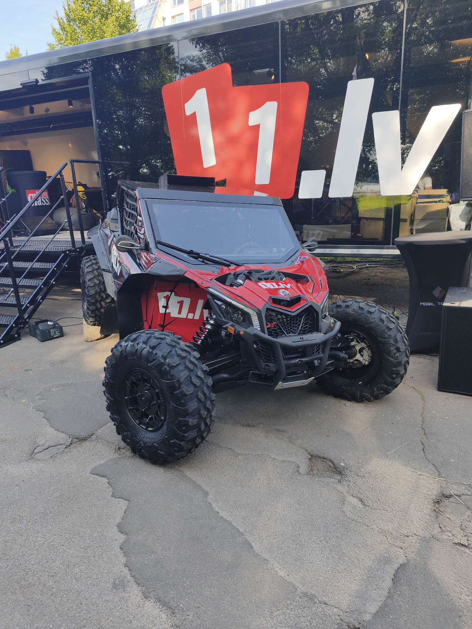 A red and black Can-Am Maverick X3 X RS side-by-side vehicle is parked on asphalt outside a large black trailer. The trailer has a large red and white logo that reads "11.lv". A black staircase leads up to the trailer's interior. To the right of the vehicle, a black fabric-covered table with a logo and a black speaker are visible. The scene appears to be in an urban outdoor environment, possibly a temporary event setup. The asphalt surface is cracked and weathered. The sky is visible in the upper portion of the image with trees and buildings in the background, indicating an outdoor setting in a city. No people are clearly depicted in a central role or engaged in significant interaction with the vehicle or the surroundings.