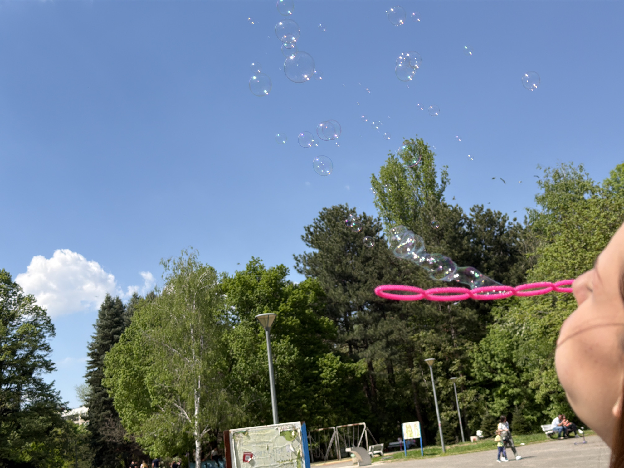 The image shows a person blowing bubbles in a park. The sky is blue with some white clouds. There are trees in the background and a lamppost to the left. In the distance, people are walking and sitting on benches. The location is likely a park in Sofia, Bulgaria, based on the presence of Cyrillic lettering on the information board.