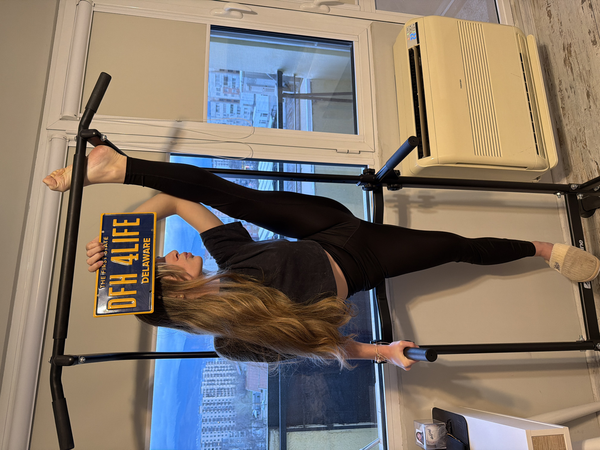 A woman is performing a split on a black pull-up bar inside an apartment. Her right leg is extended towards the right side of the frame, wearing a light-colored slipper. Her left leg is extended to the left, with her bare foot resting on the bar. She is holding a Delaware license plate reading "OFH 4 LIFE" over her head with her left hand. She is wearing black leggings and a dark crop top. Her long blonde hair is cascading down.
The pull-up bar is positioned in front of a window overlooking a city. An air conditioner is mounted in the upper right corner of the window. A partially visible object, possibly a baseball inside a display case, sits in the lower right corner. The room is illuminated by natural light. The image appears to be taken inside a residential space, not in Plovdiv, Bulgaria, but rather showing an American-themed license plate.