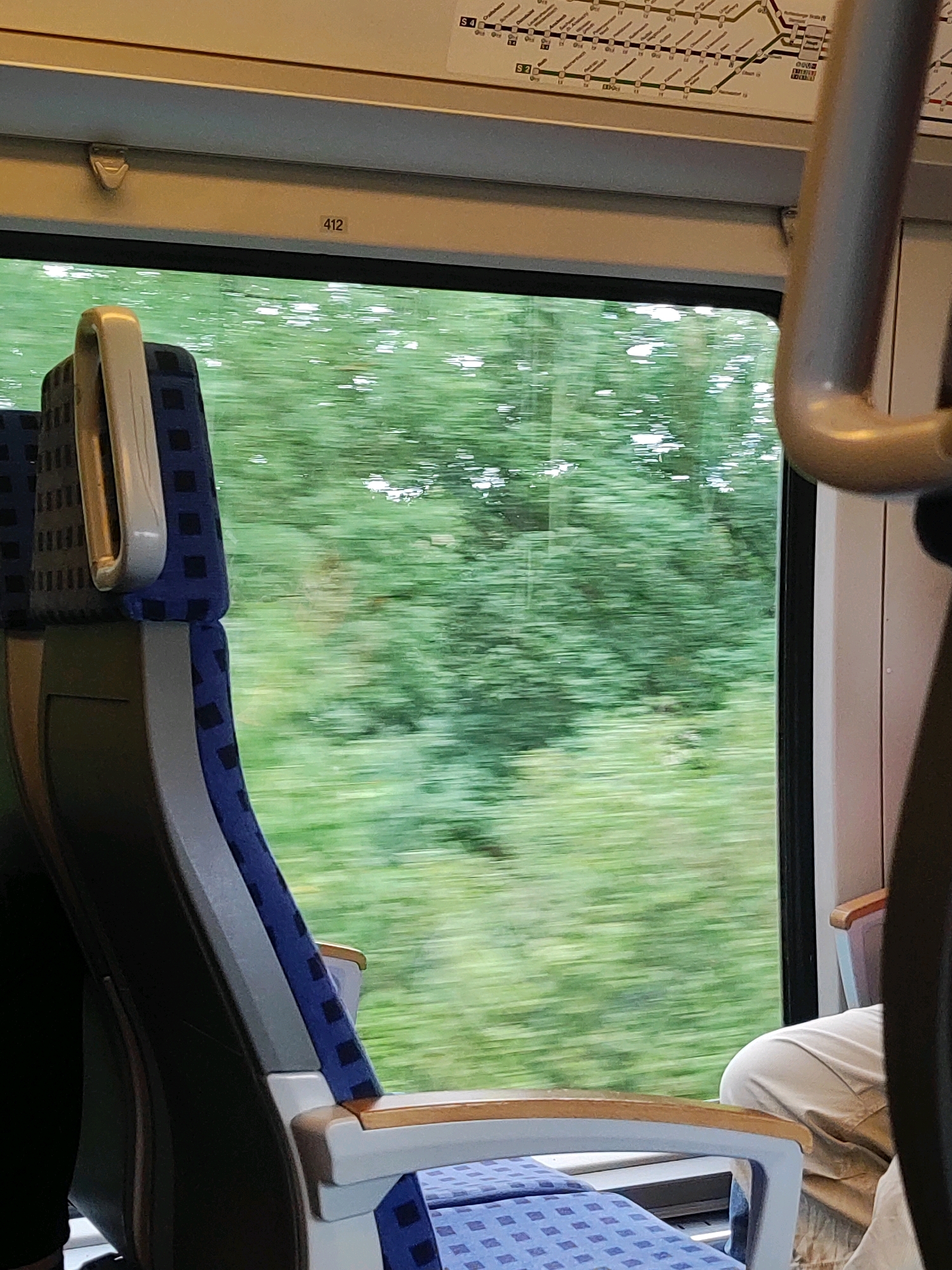 The image shows the interior of a train carriage looking out of a window. In the foreground, a blue and grey train seat with a silver handle is visible. The window shows a blurred green landscape of trees, indicating the train is in motion. Above the window, a train map or route diagram is partially visible. A small black sticker with the number "412" is affixed to the wall above the window. A person's legs, wearing light-colored trousers, are visible on the right side of the frame, seated in another part of the train. The scene is consistent with being inside a moving train in Roßtal, Germany, as implied by the route map which could display local train lines.