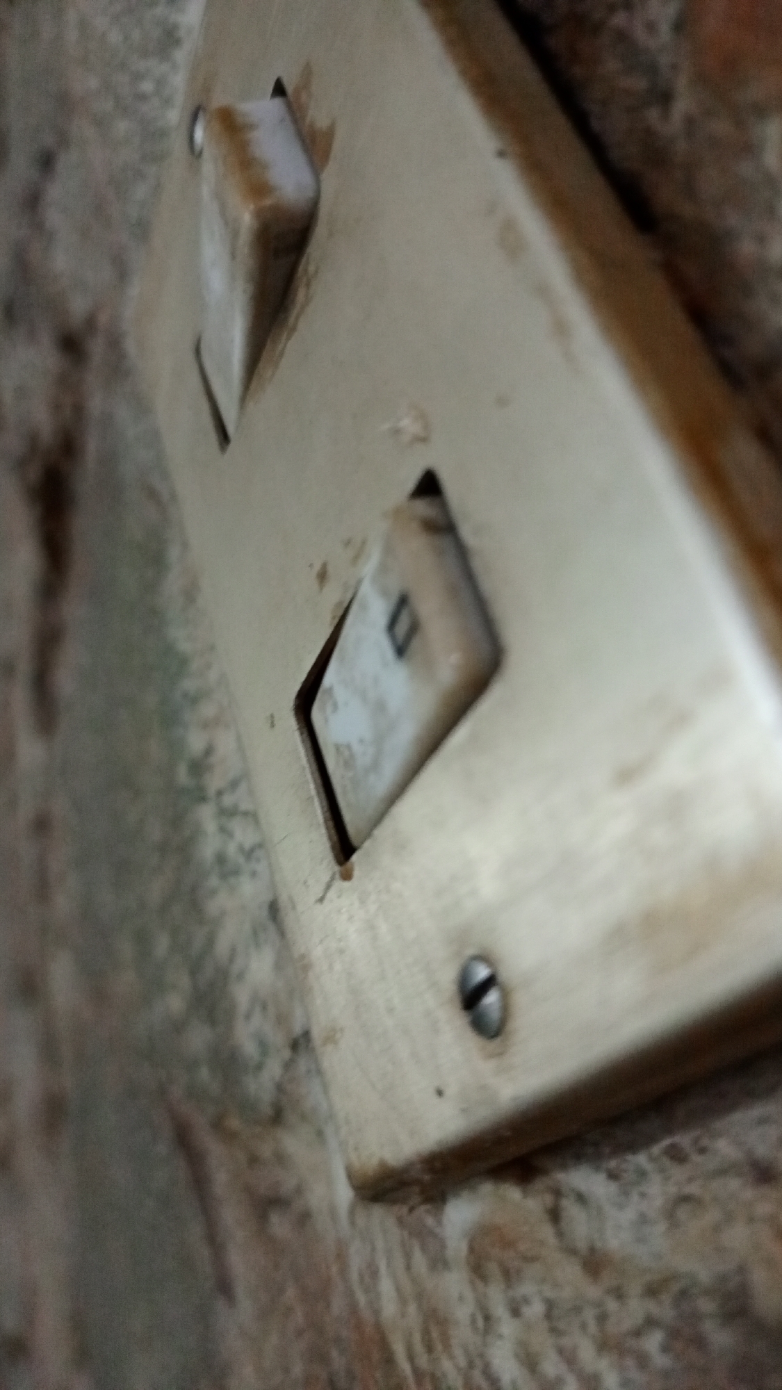 A close-up, tilted, and slightly blurred image depicts an old, discolored light switch mounted on what appears to be a rough, textured wall. The switch is cream-colored and shows signs of age and dirt accumulation. It has two toggle switches, both in the "off" or a neutral position. Two screws are visible on the lower portion of the switch plate, securing it to the wall. The wall's texture suggests it might be brick or stone. The location, Los Robles, Venezuela, is provided as background information, but there's nothing visible in the image that confirms this location.