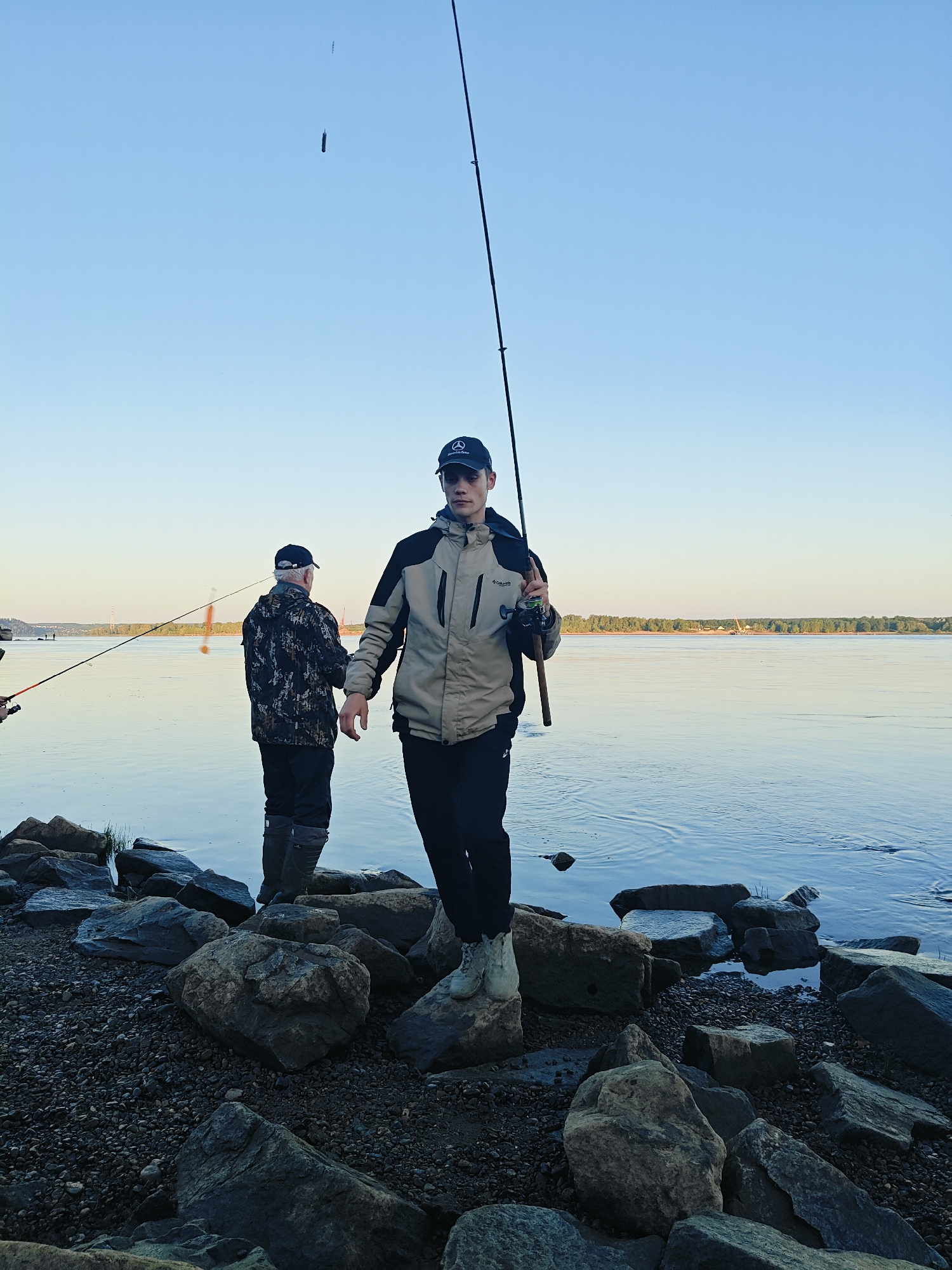 Two individuals are engaged in fishing on a rocky shoreline along a wide body of water, likely the Kama River, near the city of Chaikovsky, Russia. The foreground features a dense collection of dark, irregular rocks and gravel, transitioning to calm, blue water. In the middle ground, the two figures are positioned near the water's edge. The person closer to the camera, a young male wearing a dark baseball cap with a Mercedes-Benz logo, a beige and black jacket, and dark pants, is holding a fishing rod upright, angled towards the sky. His fishing rod has a reel attached, and the line extends upwards, with a small object, possibly bait or a lure, visible in the air. Behind him, an older individual, wearing a camouflage patterned jacket, dark pants, and high rubber boots, is also fishing, with their rod extended over the water. The background showcases a tranquil river reflecting the clear, pale blue sky. A distant shoreline with a line of trees and some structures is visible on the horizon. The lighting suggests it is either early morning or late afternoon, with soft, even illumination.