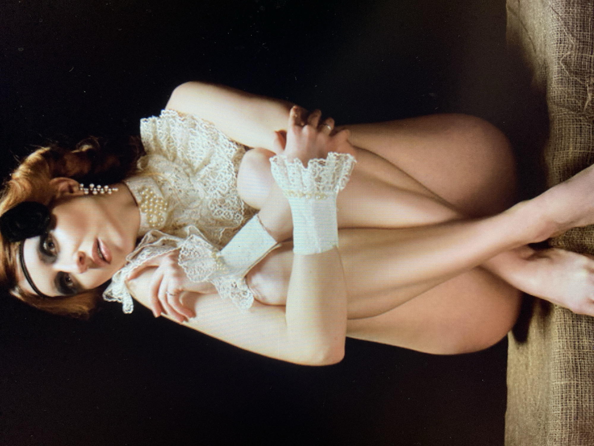A woman is sitting with her legs crossed, cradling herself. She is wearing a lace collar and cuffs and a black headband. Her eyes are looking upwards and to the left. The background is dark and undifferentiated. She is sitting on a textured surface that appears to be burlap or canvas.