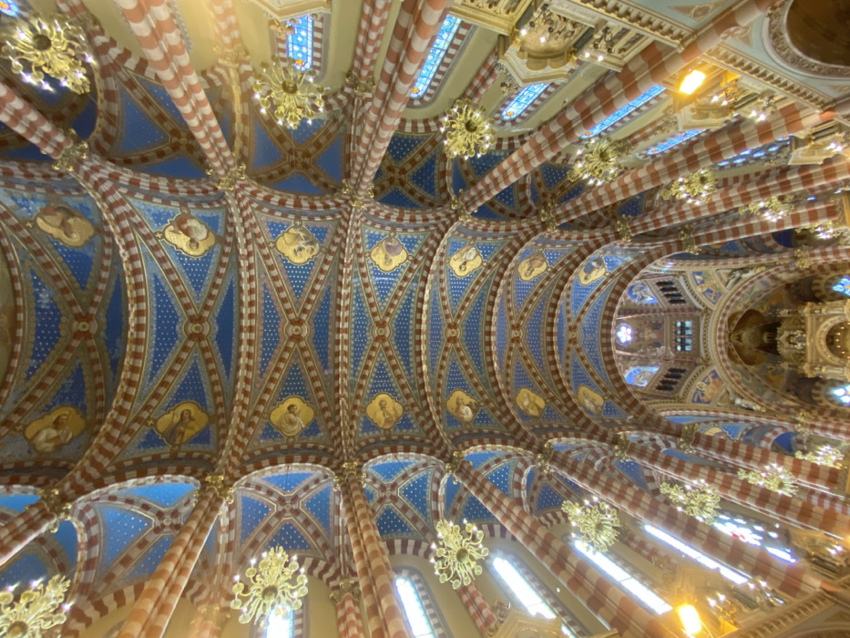 The image displays an upward-angled view of the highly ornate interior ceiling and upper architectural elements of the Basílica Santa Rosa de Lima, situated in the Monserrat neighborhood of Buenos Aires, Argentina. The vaulted ceiling is meticulously decorated with a complex geometric pattern featuring numerous deep blue segments filled with small white dots, resembling stars, alongside sections with reddish-brown and gold detailing. Gold-framed circular and oval medallions, each containing painted depictions of religious figures, are regularly spaced across the ceiling. Supporting the ceiling, prominent columns and arches are striped horizontally in alternating bands of reddish-brown and a lighter, off-white or beige color. Multiple elaborate, gold-toned chandeliers with radial arms project downwards from various points on the ceiling. Tall, arched windows with stained glass are visible along the upper side walls, allowing natural light to illuminate the interior. No individuals or active subjects are present in the scene.