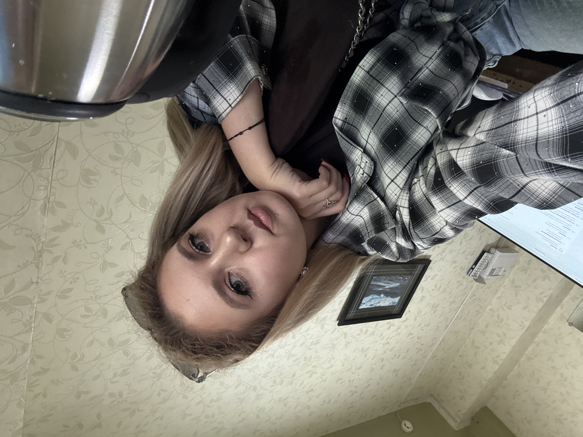 A young woman with blonde hair pulled back is taking a selfie in an office-like environment. She is wearing a black and white checkered shirt with small rhinestones and blue jeans. Her left hand is resting on her chin, and she is looking directly at the camera. In the background, there is a computer monitor, a framed picture on the wall, and a telephone. A metallic kettle is partially visible in the foreground. The walls are covered with a light-colored floral wallpaper. The overall lighting suggests it is daytime.