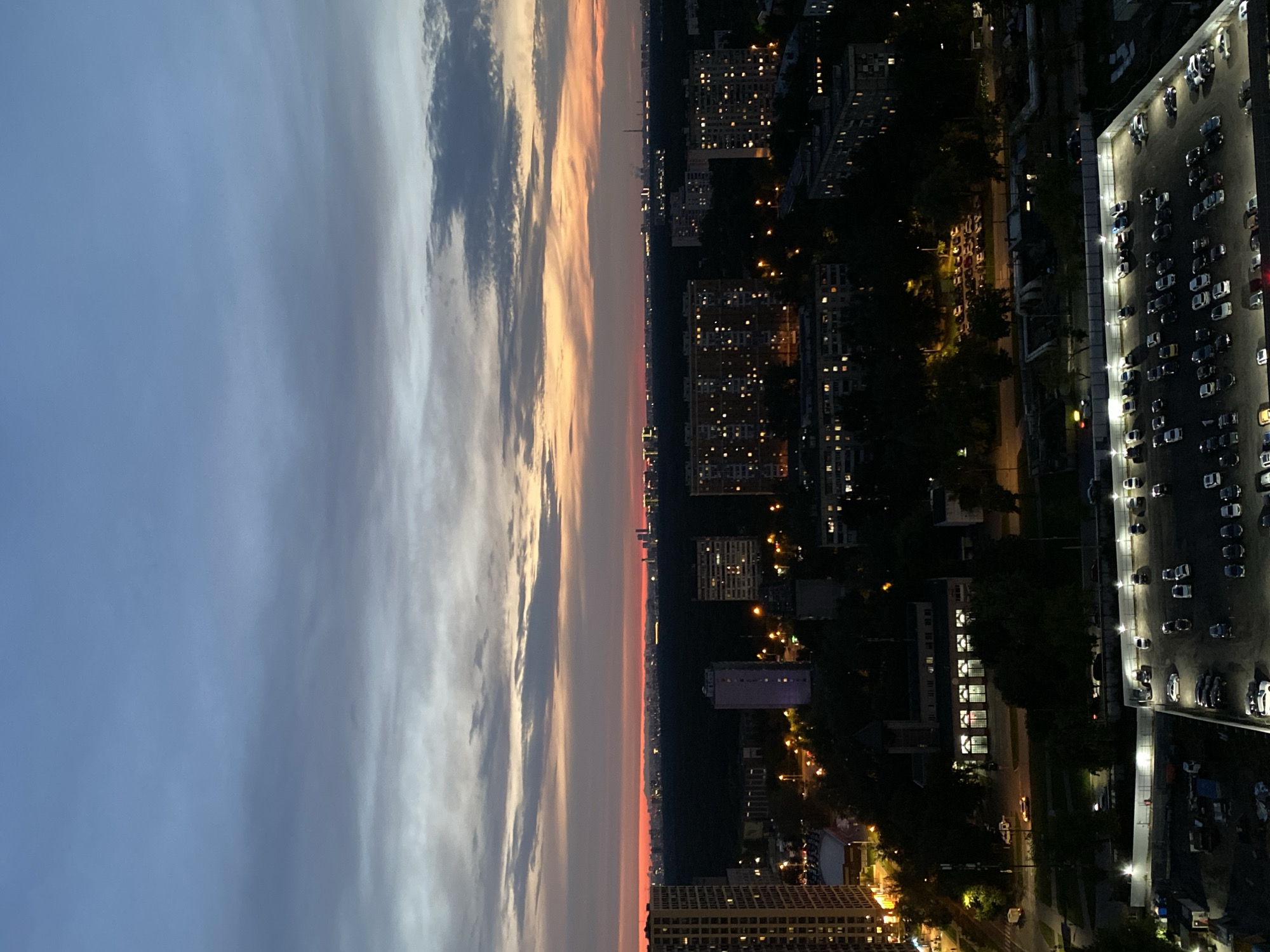 This is an aerial, dusk or twilight photograph of a sprawling urban area in Moscow, Russia. The sky is a gradient of deep blue at the top, transitioning through layers of grey clouds and ending with a band of vibrant orange and pink near the horizon, suggesting either sunrise or sunset.
The foreground is dominated by a large, well-lit outdoor parking lot filled with dozens of cars of various makes and models, parked in designated spots. The lot is bordered by a white fence and illuminated by numerous overhead lights. Adjacent to the parking lot, a multi-story building with large, illuminated windows is visible. Further into the cityscape, residential buildings of varying heights, many with lit windows, are scattered amongst dense trees. Streetlights dot the landscape, casting pools of yellow light.
In the middle ground, a line of trees separates the nearer buildings from a darker, more distant area that appears to be a forest or park. Beyond this green belt, the Moscow skyline is visible, characterized by numerous illuminated buildings, including some taller structures that suggest a city center or business district. A few industrial chimneys or towers with visible smoke or steam are also present on the distant horizon.
The overall impression is one of a densely populated urban environment at the transition from day to night, with the artificial lights of the city beginning to dominate the natural twilight. No people are clearly discernible in the image.